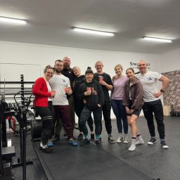 Akademia Sprawności - Grupa dziewięciu osób, prawdopodobnie uczestników treningu personalnego, pozuje z uśmiechem w siłowni z sprzętem do ćwiczeń siłowych w tle, trzymając kubki z napojami.