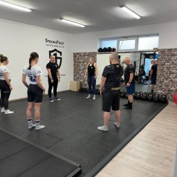 Akademia Sprawności - Grupa osób w sali gimnastycznej, część z nich ubrana w koszulki z logo, ustawiona w kręgu, prawdopodobnie podczas instruktażu lub rozgrzewki, w tle widoczne kettlebells i logo StrongFirst.