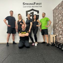 Akademia Sprawności - Grupa osób w sportowych strojach pozuje w Kłodzku w siłowni z napisem StrongFirst Certified Instructor na ścianie, obok ceglanej ściany i sprzętu do ćwiczeń.