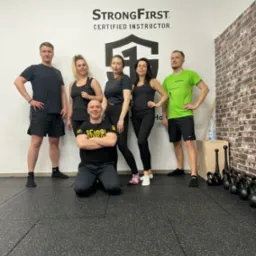 Grupa osób w sportowych strojach pozuje w Kłodzku w siłowni z napisem StrongFirst Certified Instructor na ścianie, obok ceglanej ściany i sprzętu do ćwiczeń.