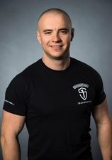 Portret uśmiechniętego mężczyzny o ogolonej głowie w czarnej koszulce z logo szkoły walki na szarym tle.