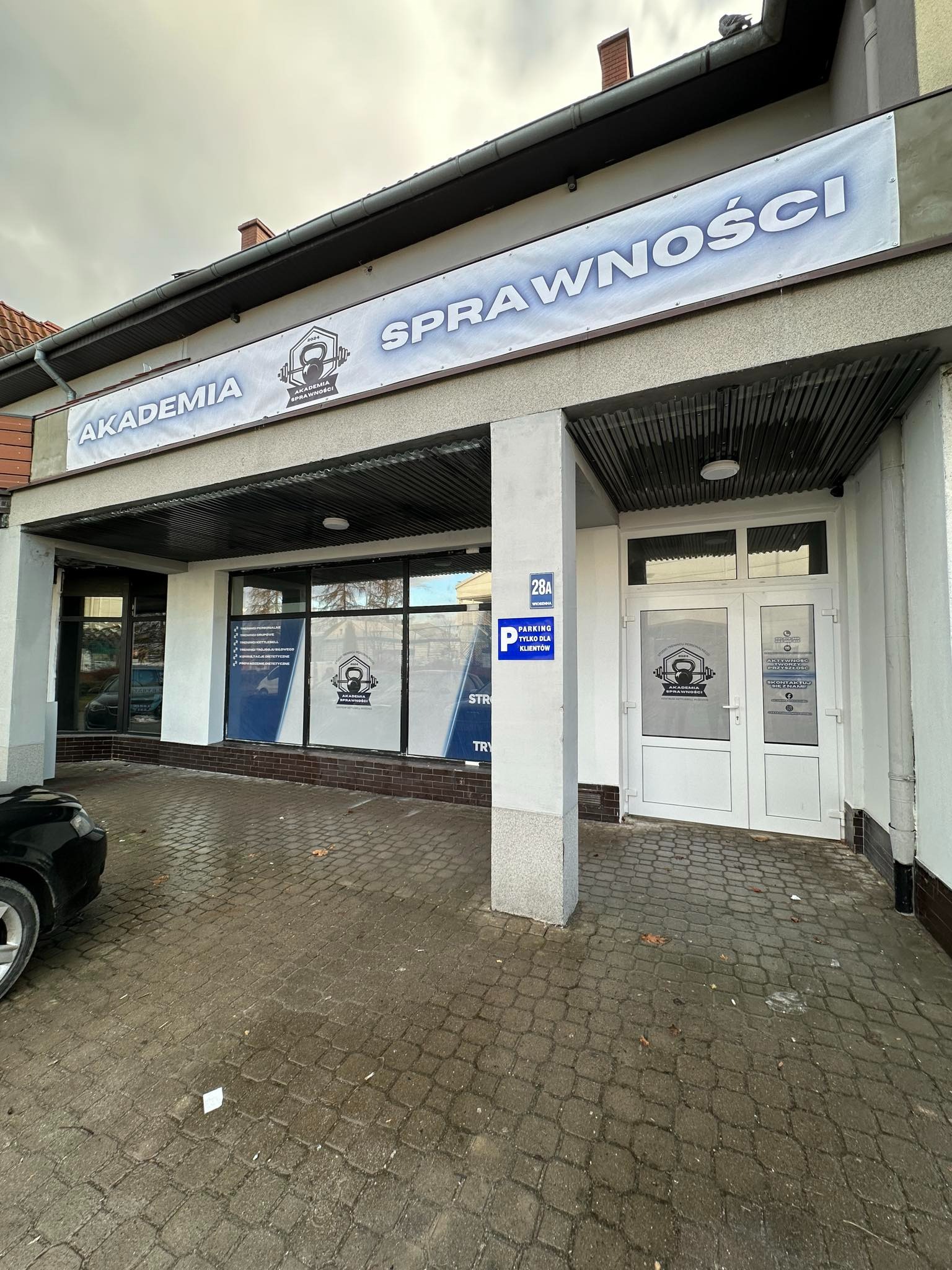 Fasada budynku Akademii Sprawności z szyldem, oknami z informacjami o treningach i logo, tabliczką z adresem oraz informacją o parkingu dla klientów.