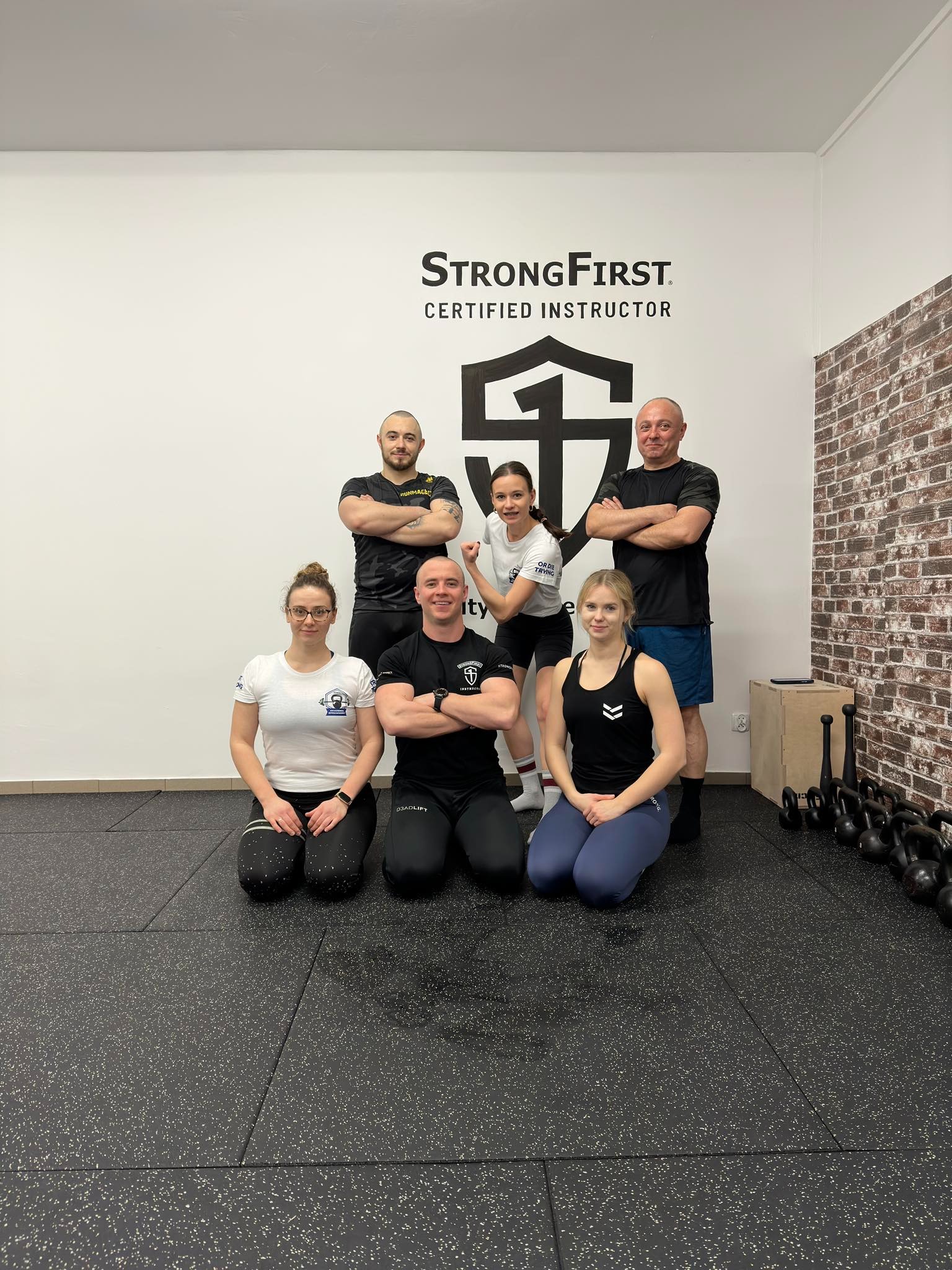 Grupa sześciu osób w strojach sportowych pozuje w siłowni na tle ściany z logo StrongFirst Certified Instructor, obok widoczne kettlebells.