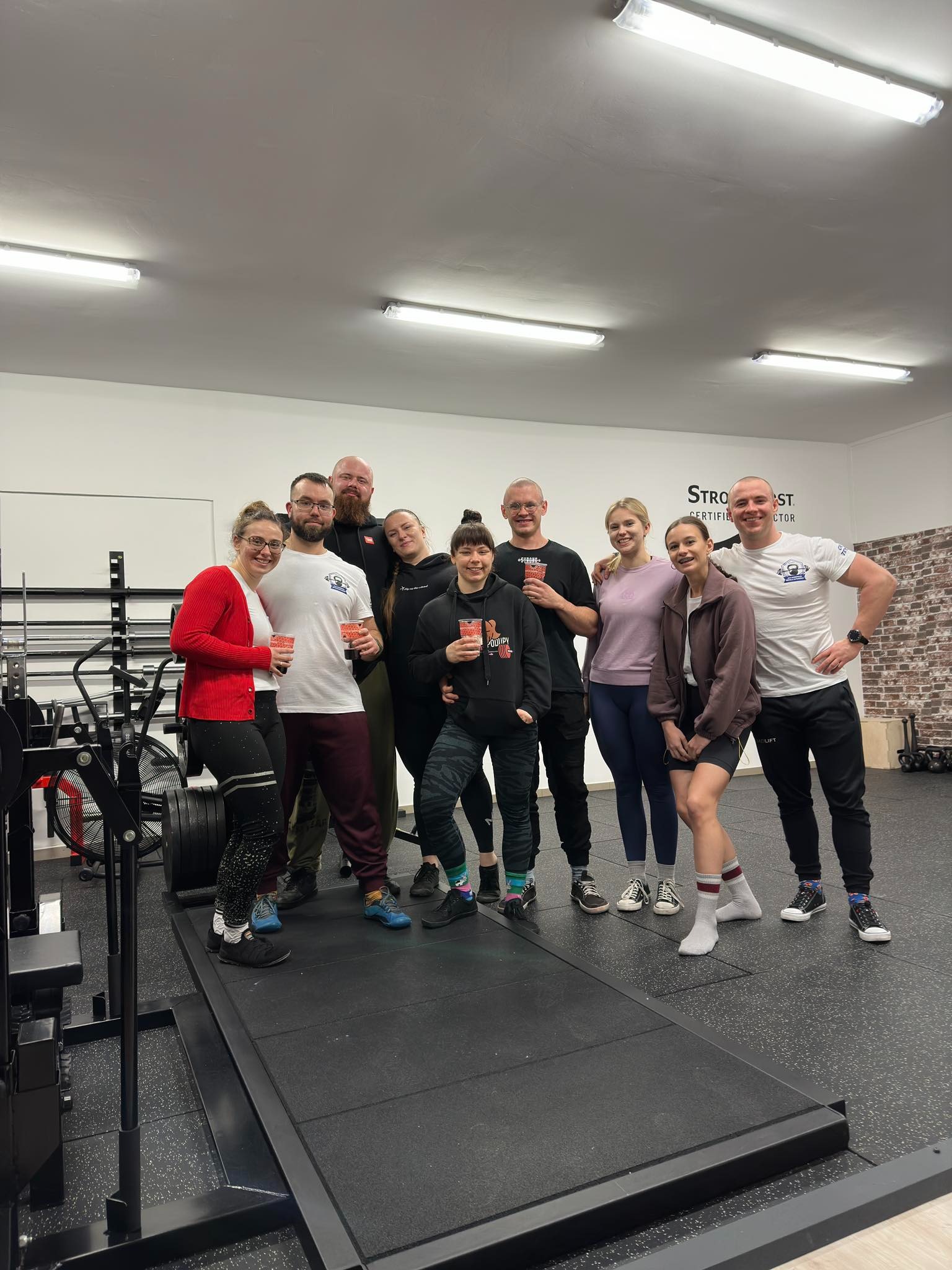Grupa dziewięciu osób, prawdopodobnie uczestników treningu personalnego, pozuje z uśmiechem w siłowni z sprzętem do ćwiczeń siłowych w tle, trzymając kubki z napojami.