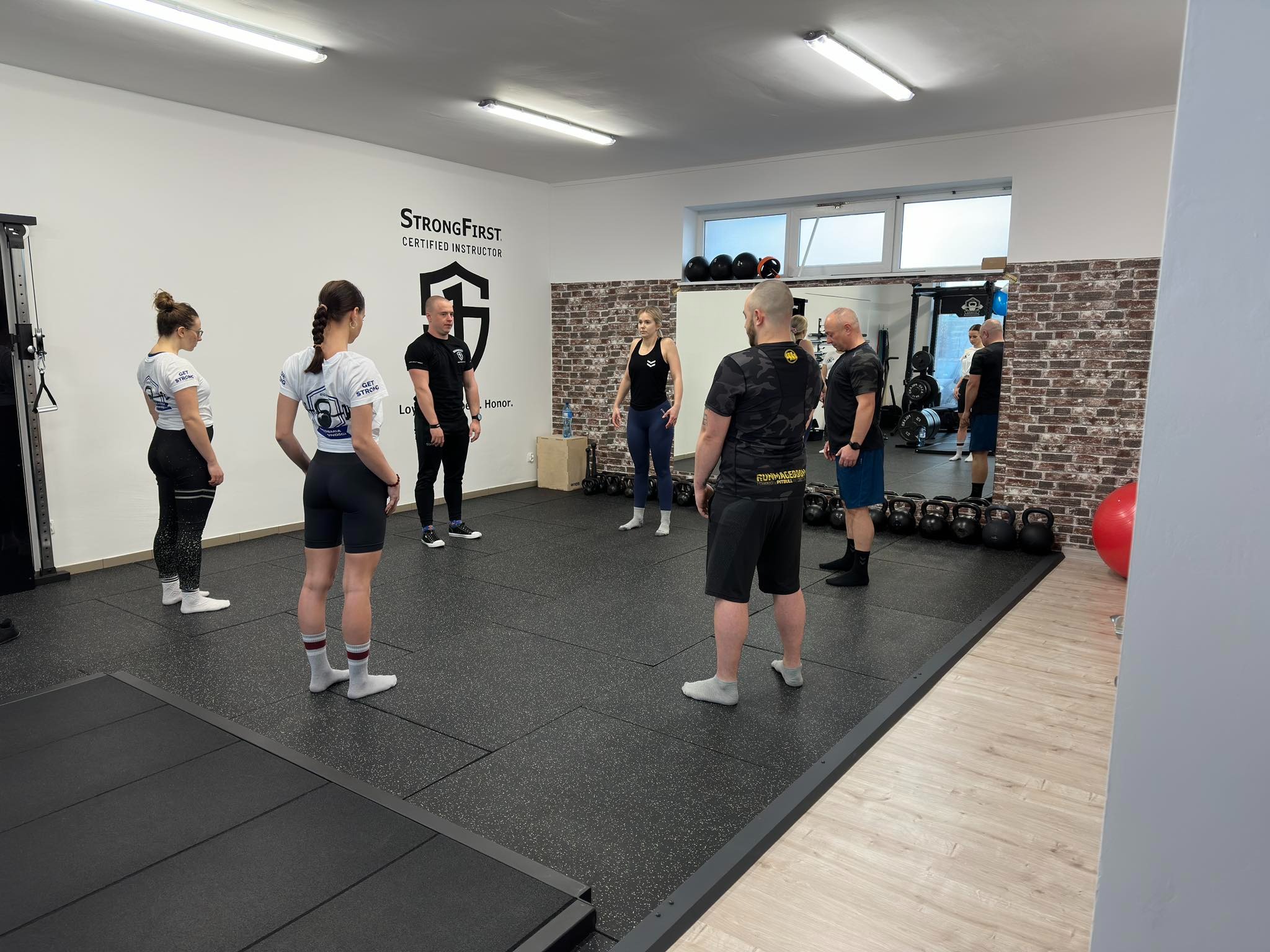 Grupa osób w sali gimnastycznej, część z nich ubrana w koszulki z logo, ustawiona w kręgu, prawdopodobnie podczas instruktażu lub rozgrzewki, w tle widoczne kettlebells i logo StrongFirst.
