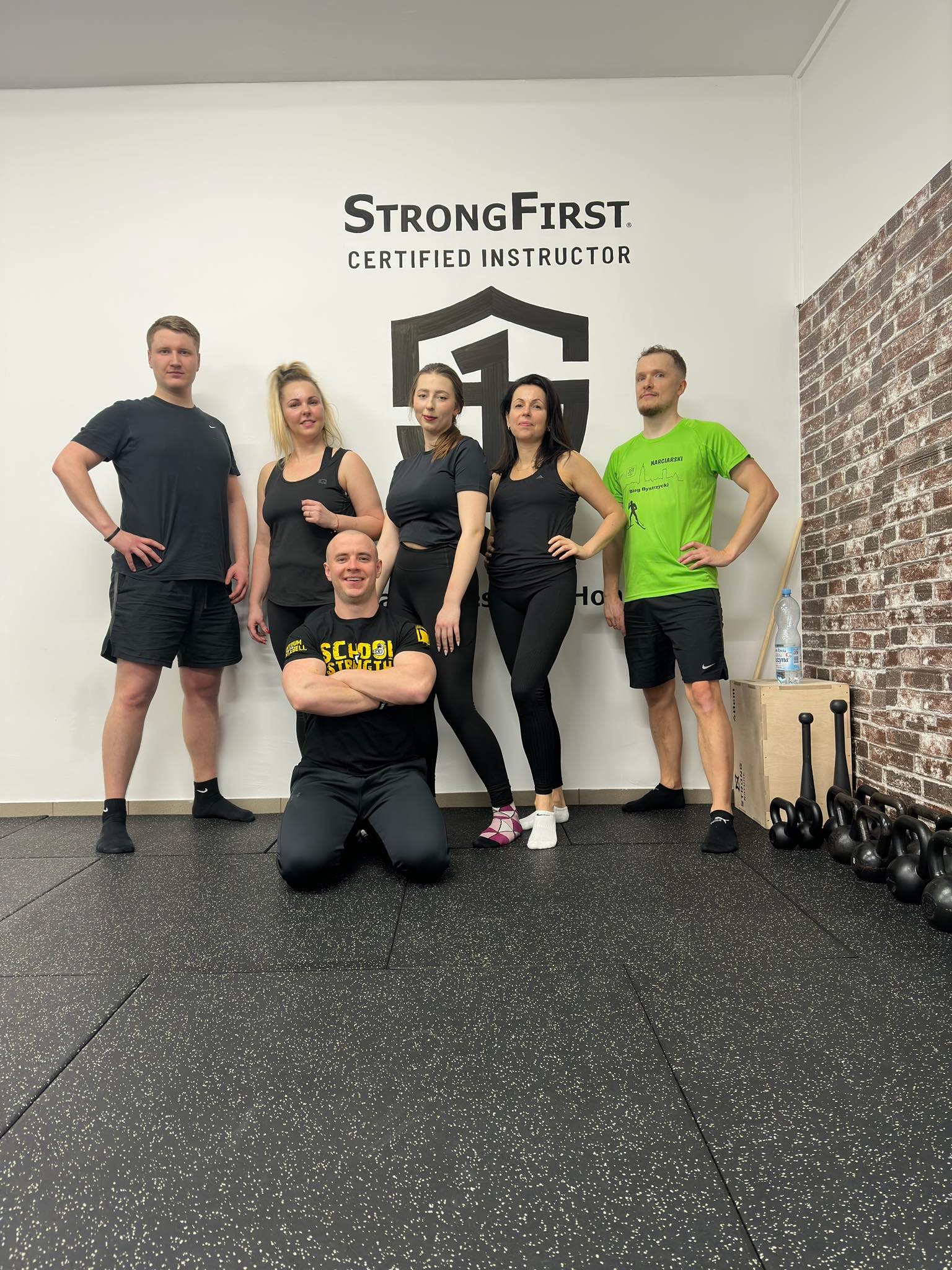 Grupa osób w sportowych strojach pozuje w Kłodzku w siłowni z napisem StrongFirst Certified Instructor na ścianie, obok ceglanej ściany i sprzętu do ćwiczeń.