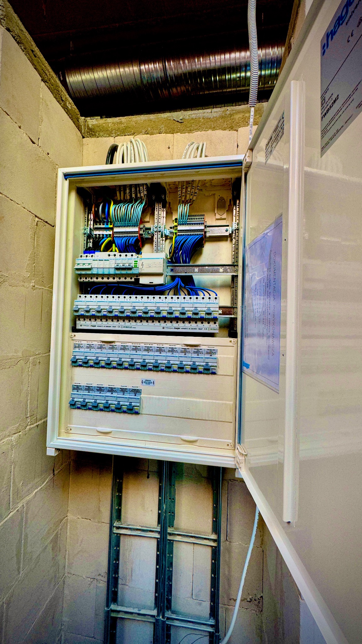 Otwarta skrzynka elektryczna Hager z widocznymi bezpiecznikami, przewodami i dokumentacją powykonawczą na ścianie z bloków. Widoczne kable i rury instalacyjne.