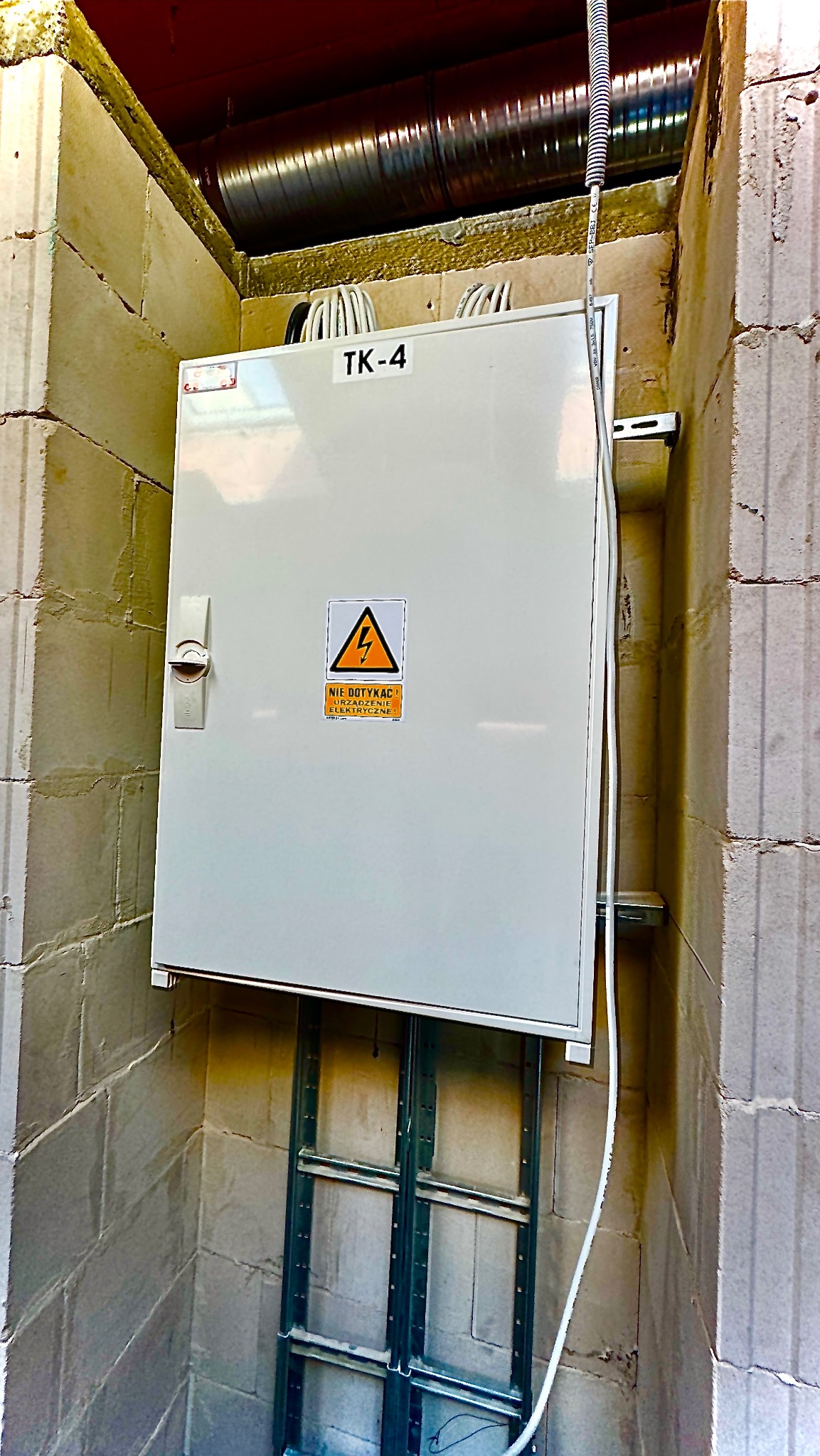 Biała skrzynka elektryczna TK-4 z ostrzeżeniem 'Nie dotykać' na surowej ścianie z betonu komórkowego. Widoczne kable i rura wentylacyjna pod sufitem.