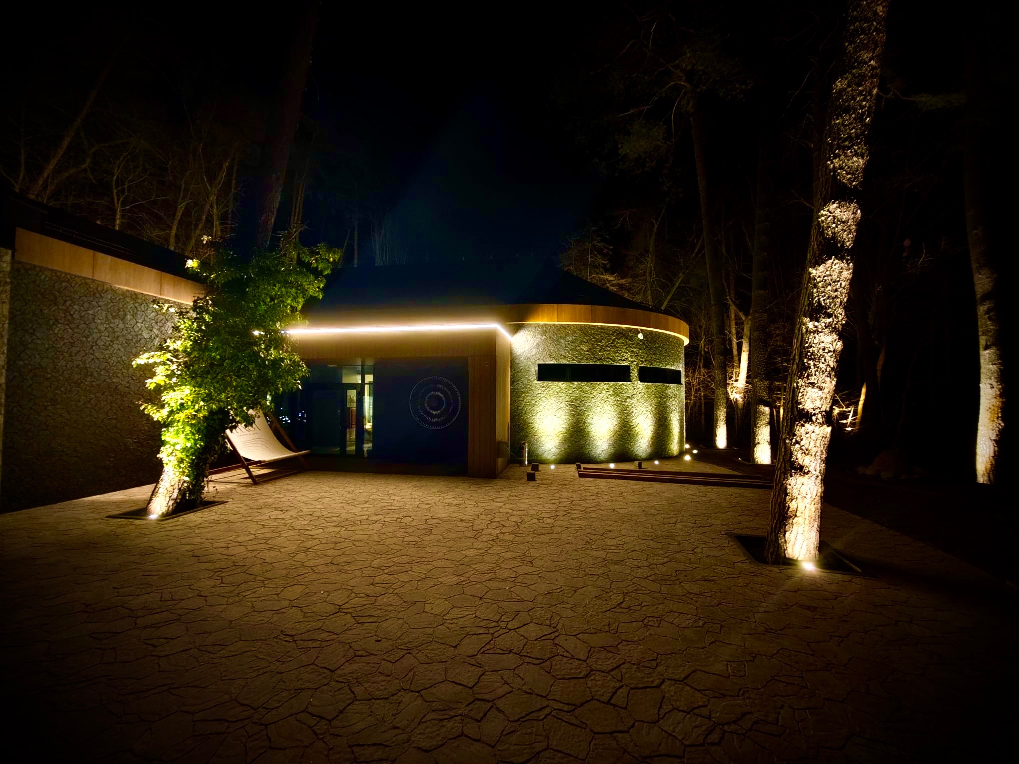 Nocne oświetlenie budynku z kamienia i drewna, z podświetlonymi drzewami i brukowaną ścieżką, tworzące ciepłą, zapraszającą atmosferę.