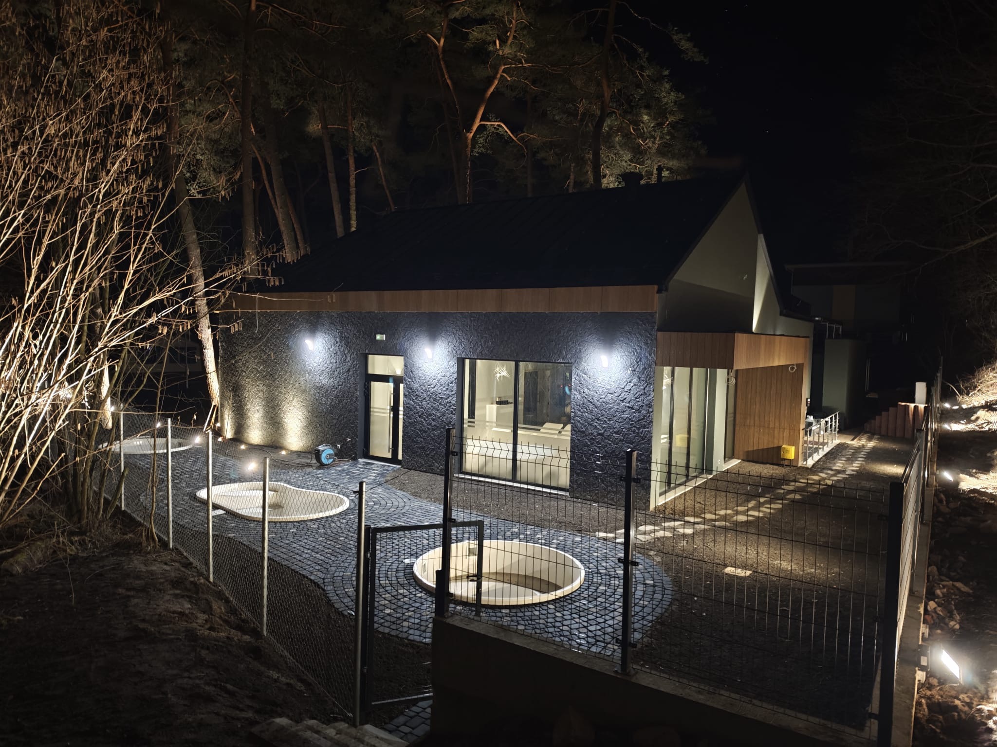 Nowoczesny dom z podświetlonym ogrodem nocą. Widoczne dwa okrągłe, wbudowane baseny otoczone kostką brukową i metalowym ogrodzeniem. Oświetlenie elewacji.