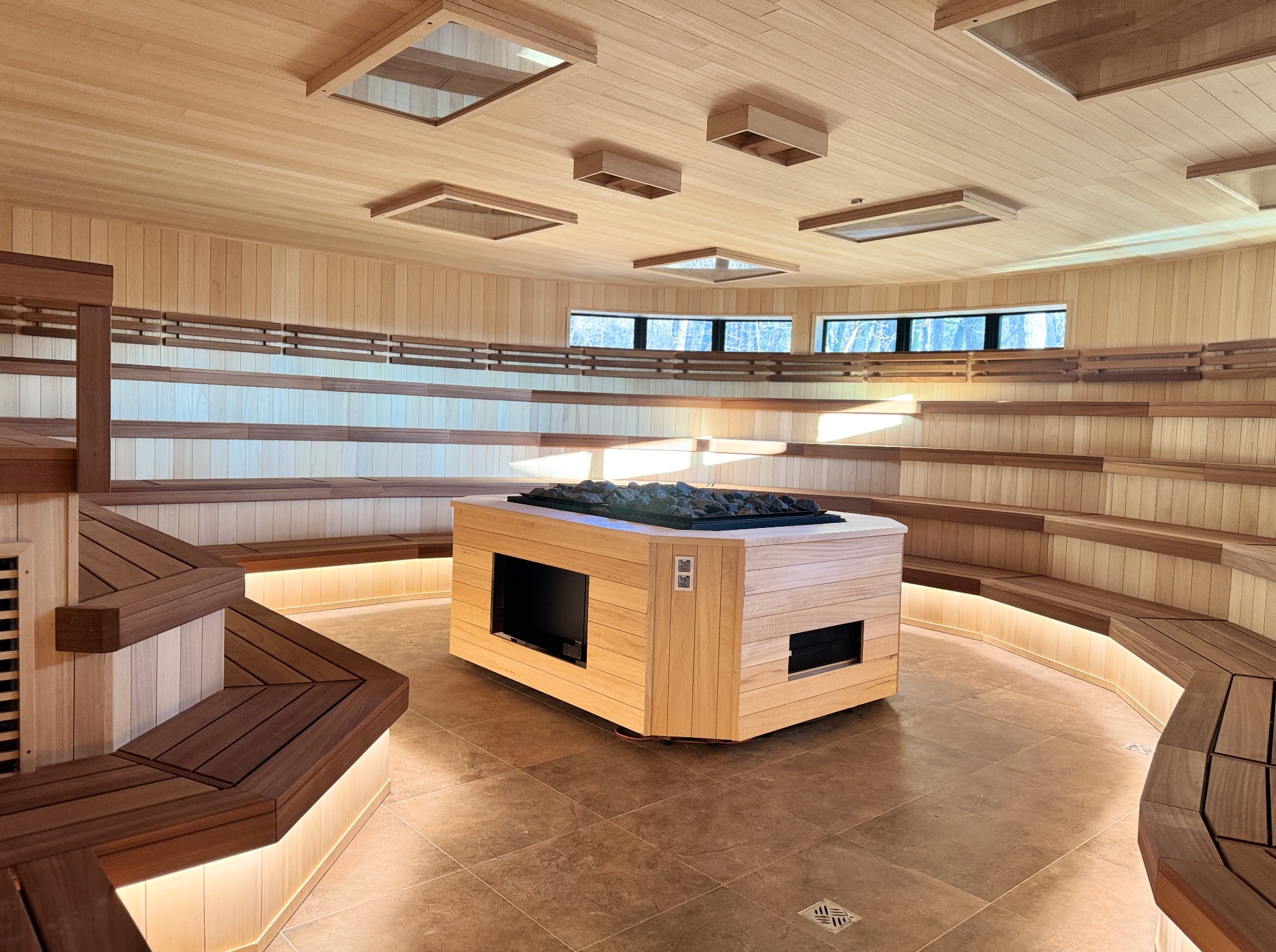 Jasne drewno w saunie: ławy na różnych poziomach, centralnie piec z kamieniami, okna z widokiem na zewnątrz, podświetlenie LED. Nowoczesny design i relaksująca atmosfera.