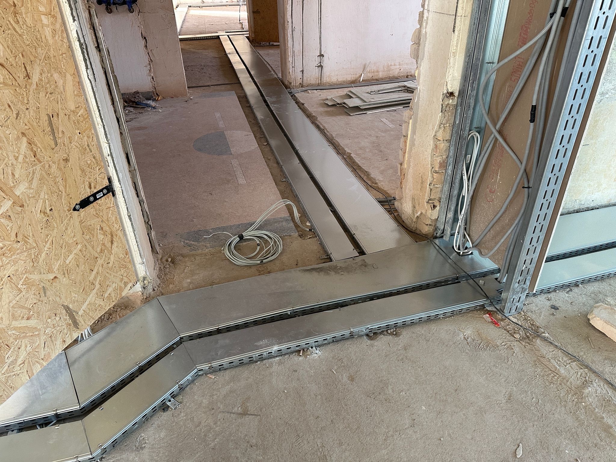 Metalowe korytka kablowe ułożone wzdłuż podłogi w surowym wnętrzu. Widoczne przewody i elementy konstrukcyjne ścian. Korytka prowadzone wzdłuż ścian i przez otwory drzwiowe.
