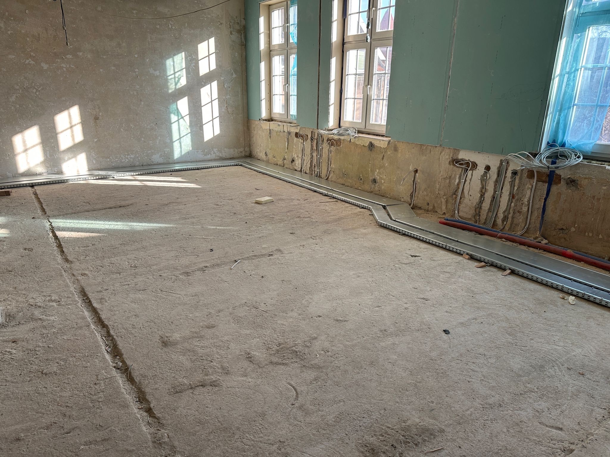 Wnętrze w trakcie remontu: koryta kablowe ułożone wzdłuż ścian, wystające przewody elektryczne, ślady po instalacjach w ścianach, surowa podłoga betonowa.