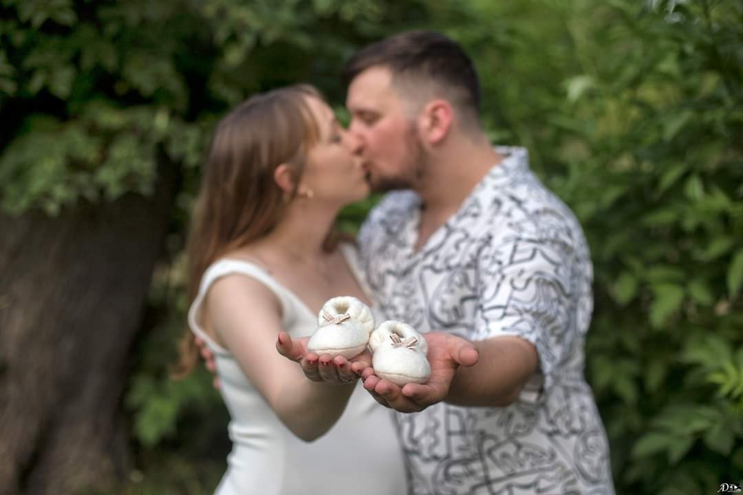 Kobieta w białej sukience i mężczyzna w koszuli w abstrakcyjne wzory całują się, trzymając w dłoniach białe, filcowe buciki niemowlęce z kokardkami na tle zieleni.