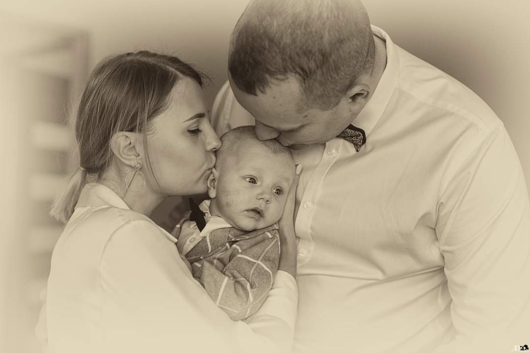 Rodzice całują niemowlę w głowę i policzek w ciepłych odcieniach sepii; ojciec w białej koszuli z muszką, matka w białej koszuli i kolczykach, dziecko w koszuli w kratę i z muchą.