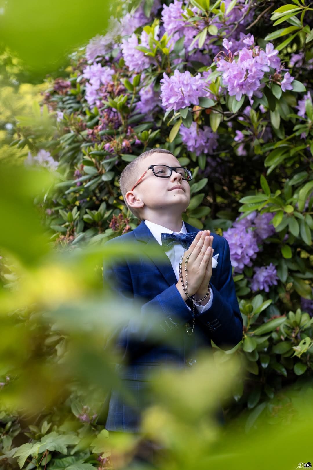 Chłopiec w garniturze z różańcem, spoglądający w górę, na tle kwitnących rododendronów. Ujęcie z dołu, z zielonymi liśćmi na pierwszym planie.