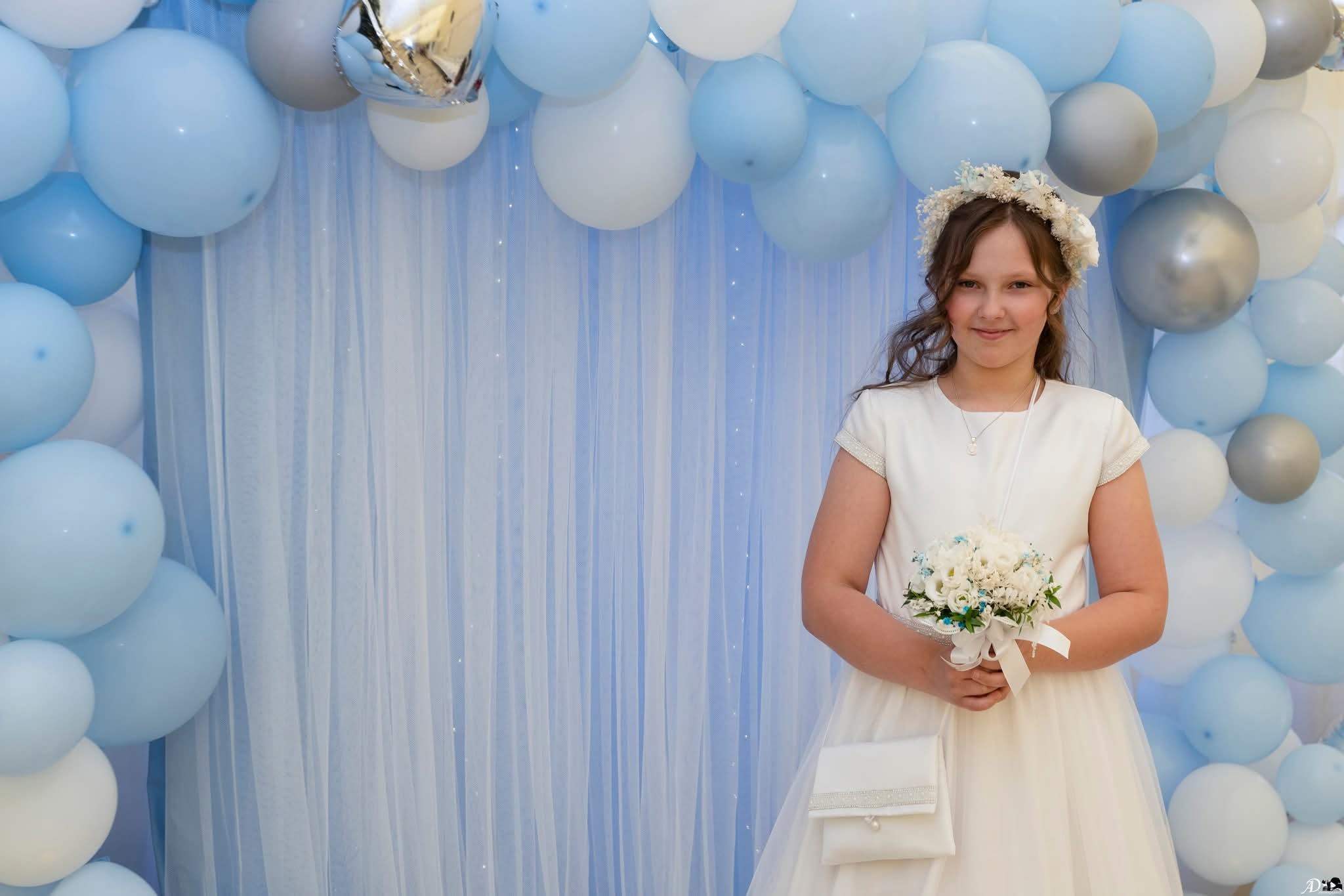 Dziewczynka w białej sukience komunijnej z wiankiem na głowie i bukietem w rękach, na tle dekoracji z błękitnych i białych balonów oraz tiulowej zasłony.