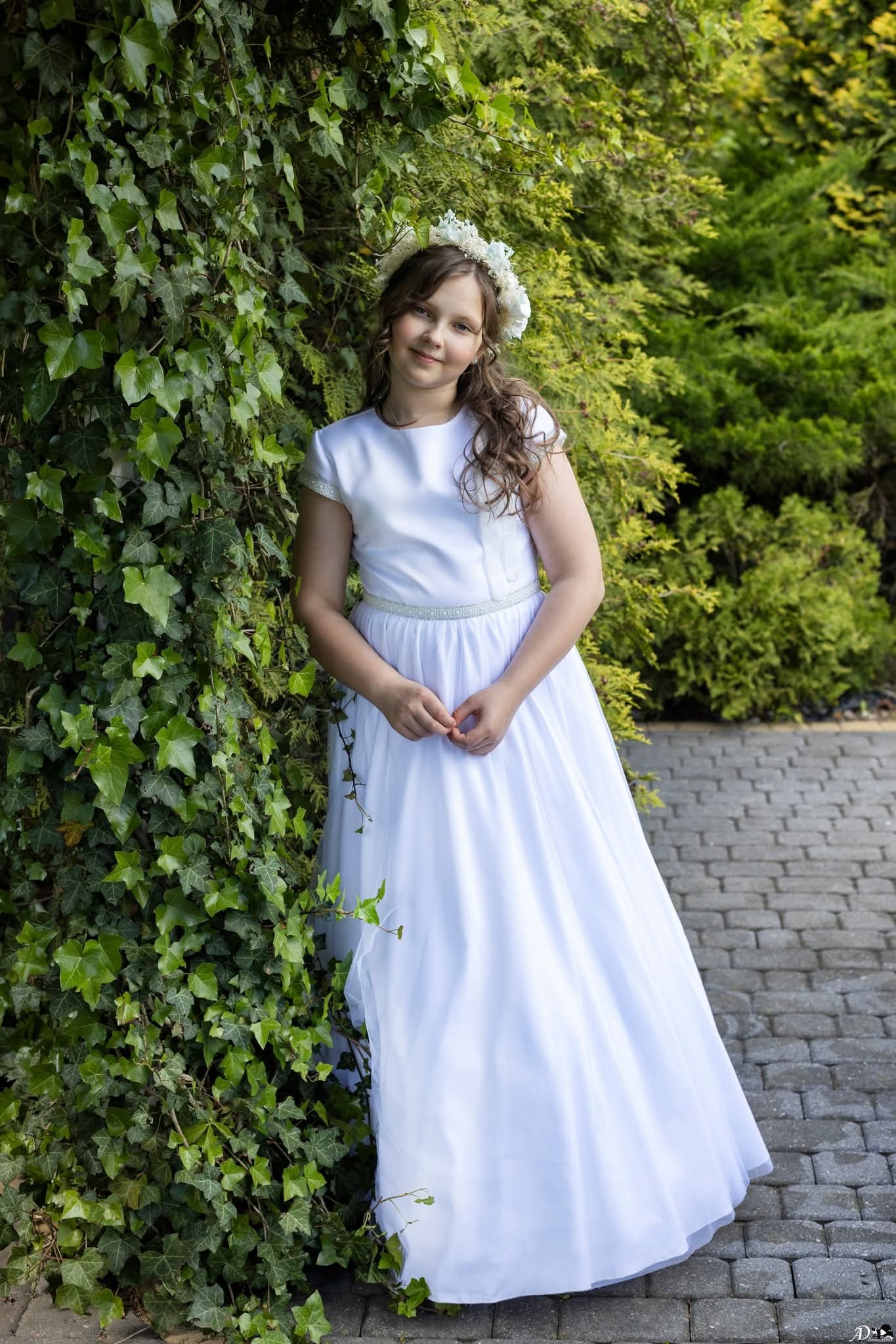 Dziewczynka w białej sukience komunijnej, z wiankiem na głowie, pozuje oparta o ścianę z bluszczu na tle zieleni.