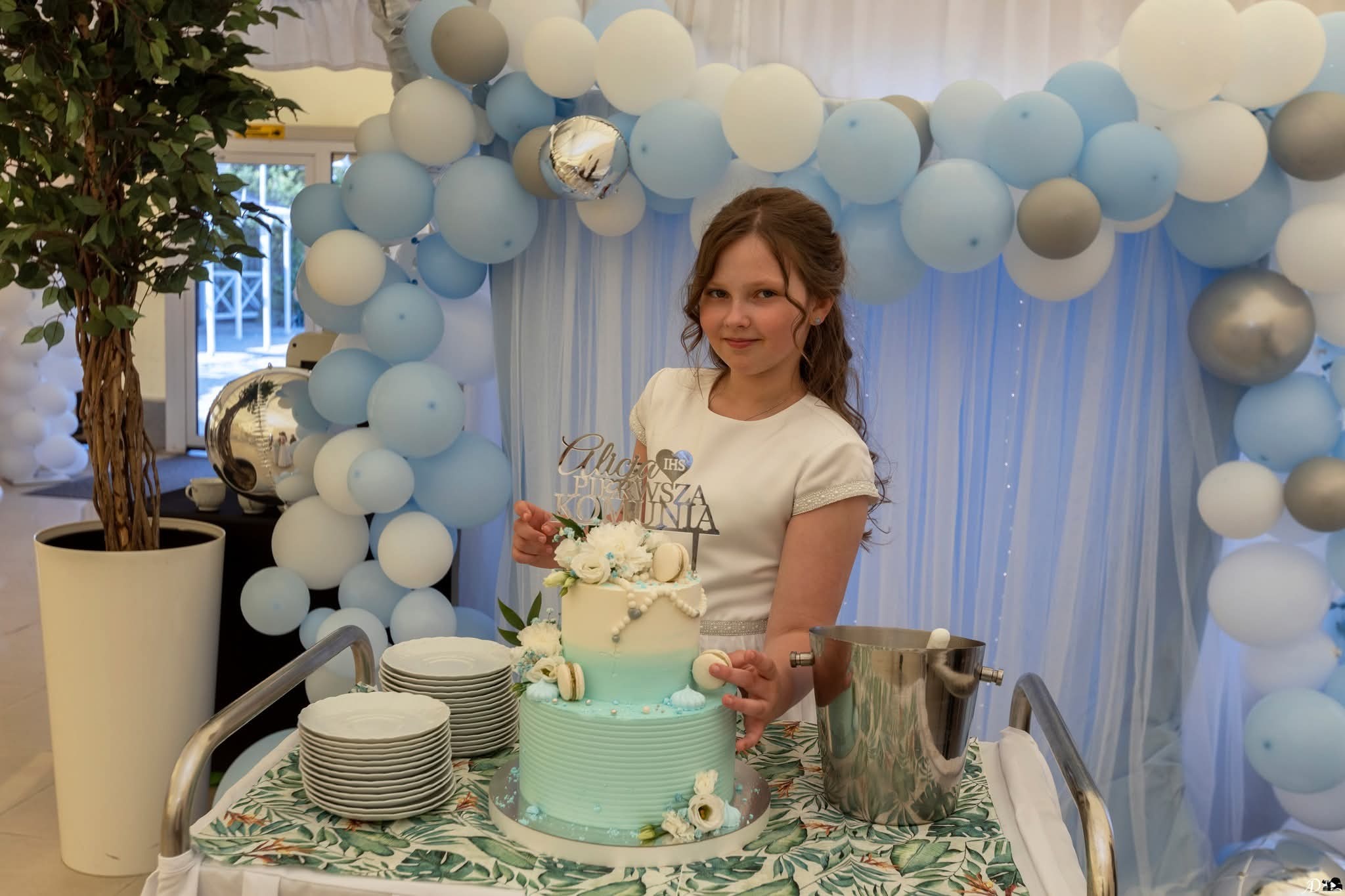 Dziewczynka w białej sukience prezentuje tort komunijny na wózku, otoczona dekoracjami z balonów w odcieniach błękitu i bieli. Uśmiechnięta, z widocznym napisem 'Pierwsza Komunia'.