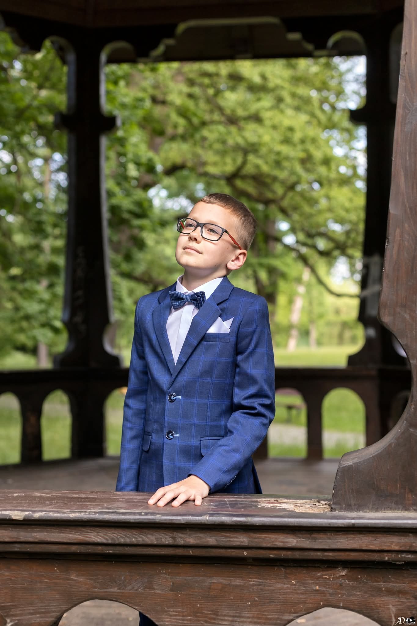 Chłopiec w niebieskim garniturze w kratę, muszka, okulary, patrzy w górę, drewniana altana w tle, sesja plenerowa, delikatne światło.