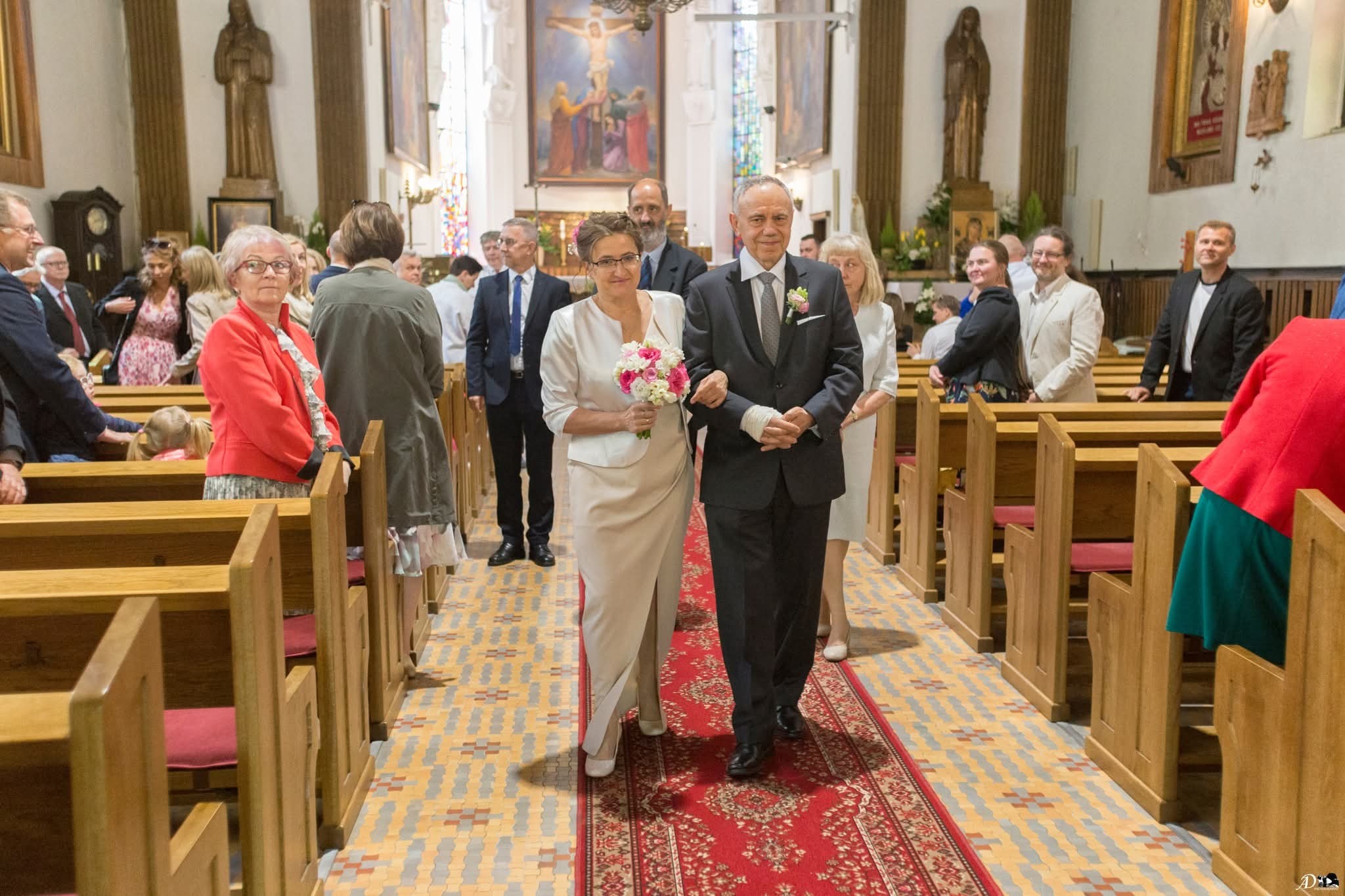 Para młoda, idąca w kościele po czerwonym dywanie, otoczona gośćmi. Panna młoda trzyma bukiet. W tle widoczne obrazy i figury świętych.