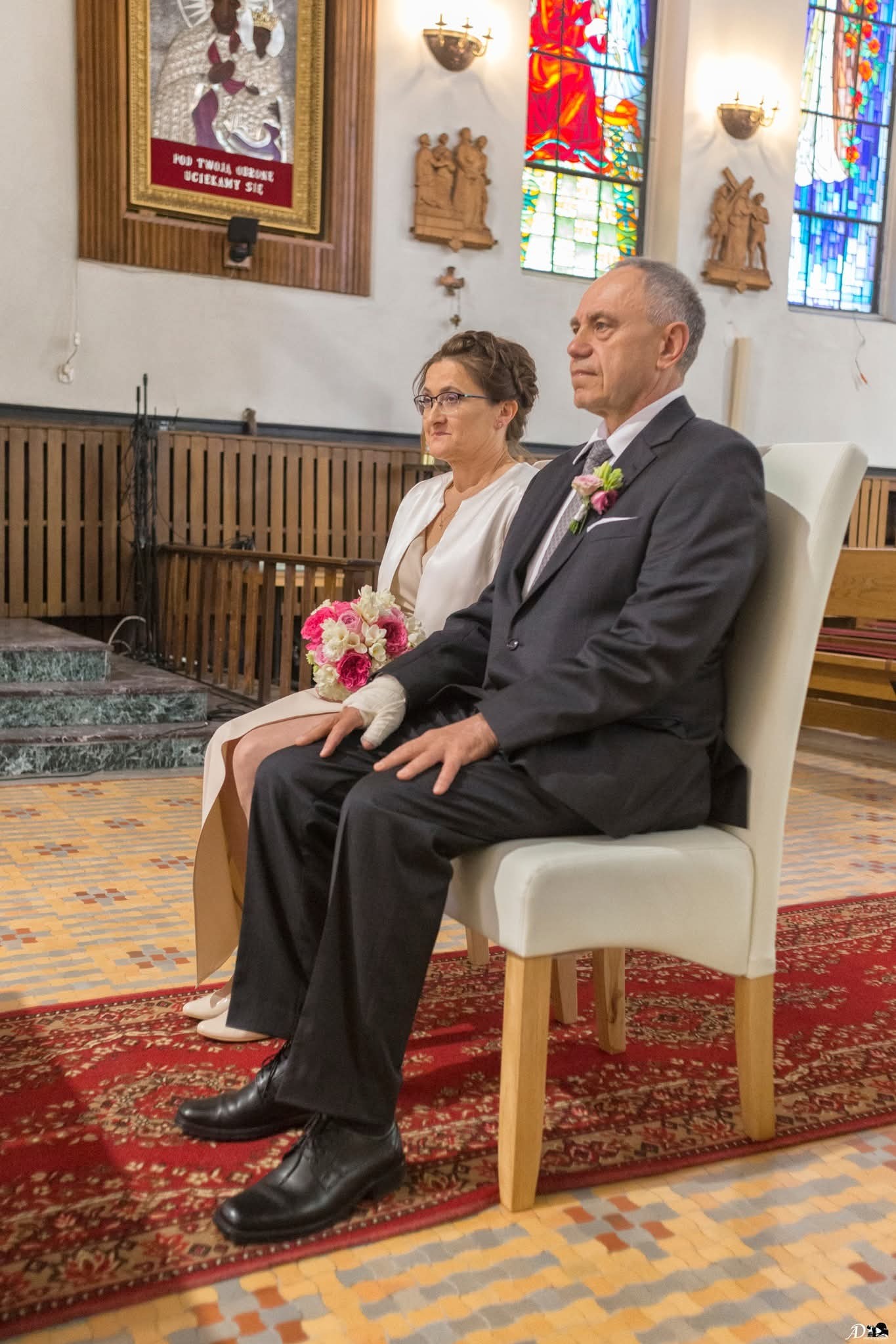 Para w kościele siedzi na krzesłach podczas ceremonii ślubnej, w tle witraże i obraz religijny. Bukiet kwiatów na kolanach panny młodej.