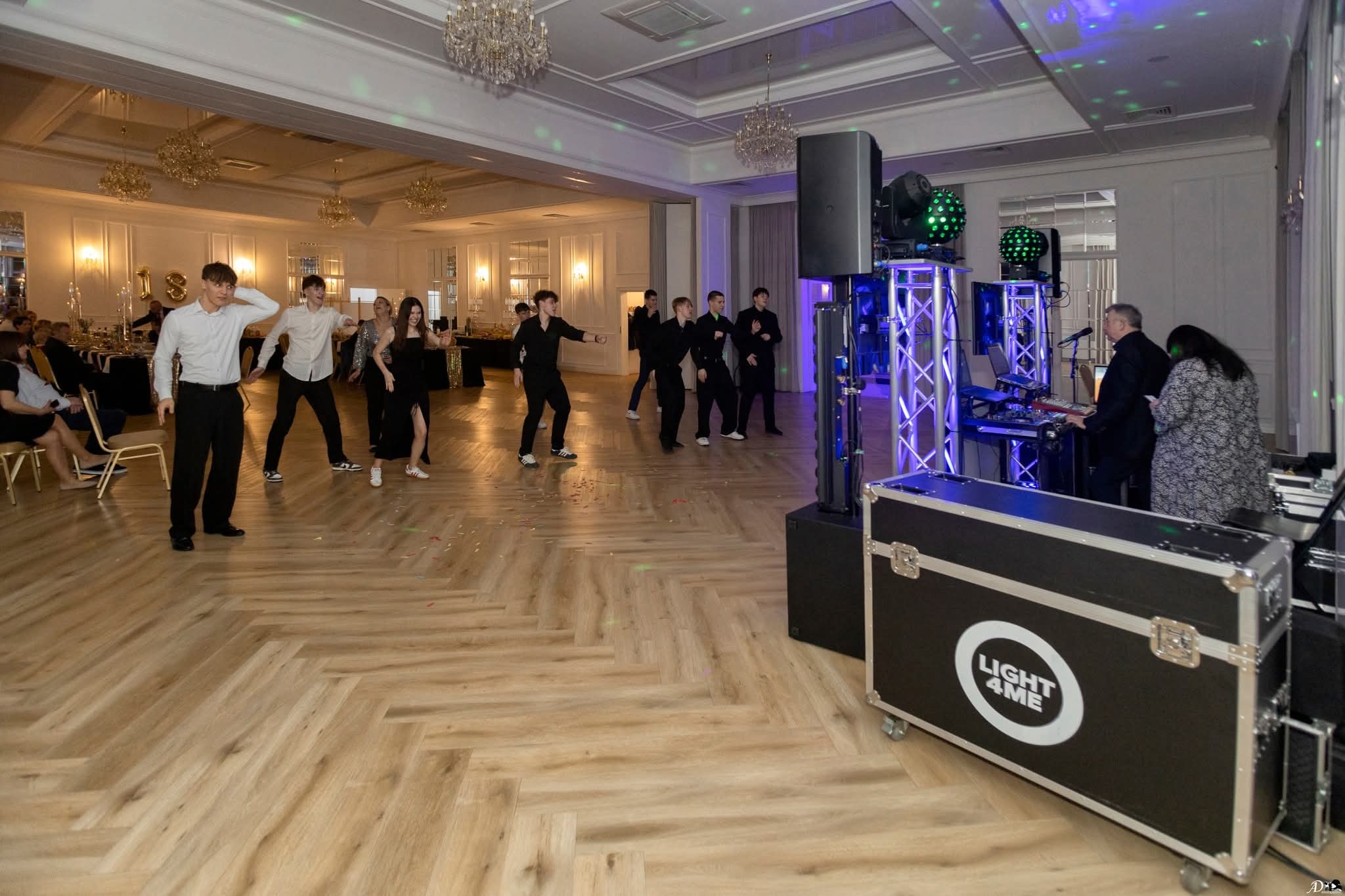 Sala balowa z parkietem w jodełkę, oświetleniem dyskotekowym i grupą tańczących osób podczas imprezy. Na pierwszym planie sprzęt nagłośnieniowy.