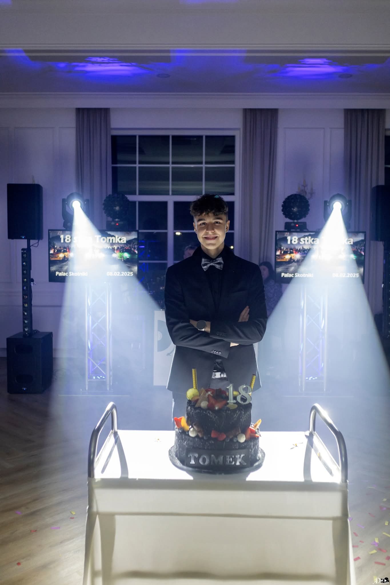 Młody mężczyzna w garniturze pozuje z tortem urodzinowym 'Tomek' na tle dekoracji świetlnych i ekranów z napisem '18-stka Tomka'.