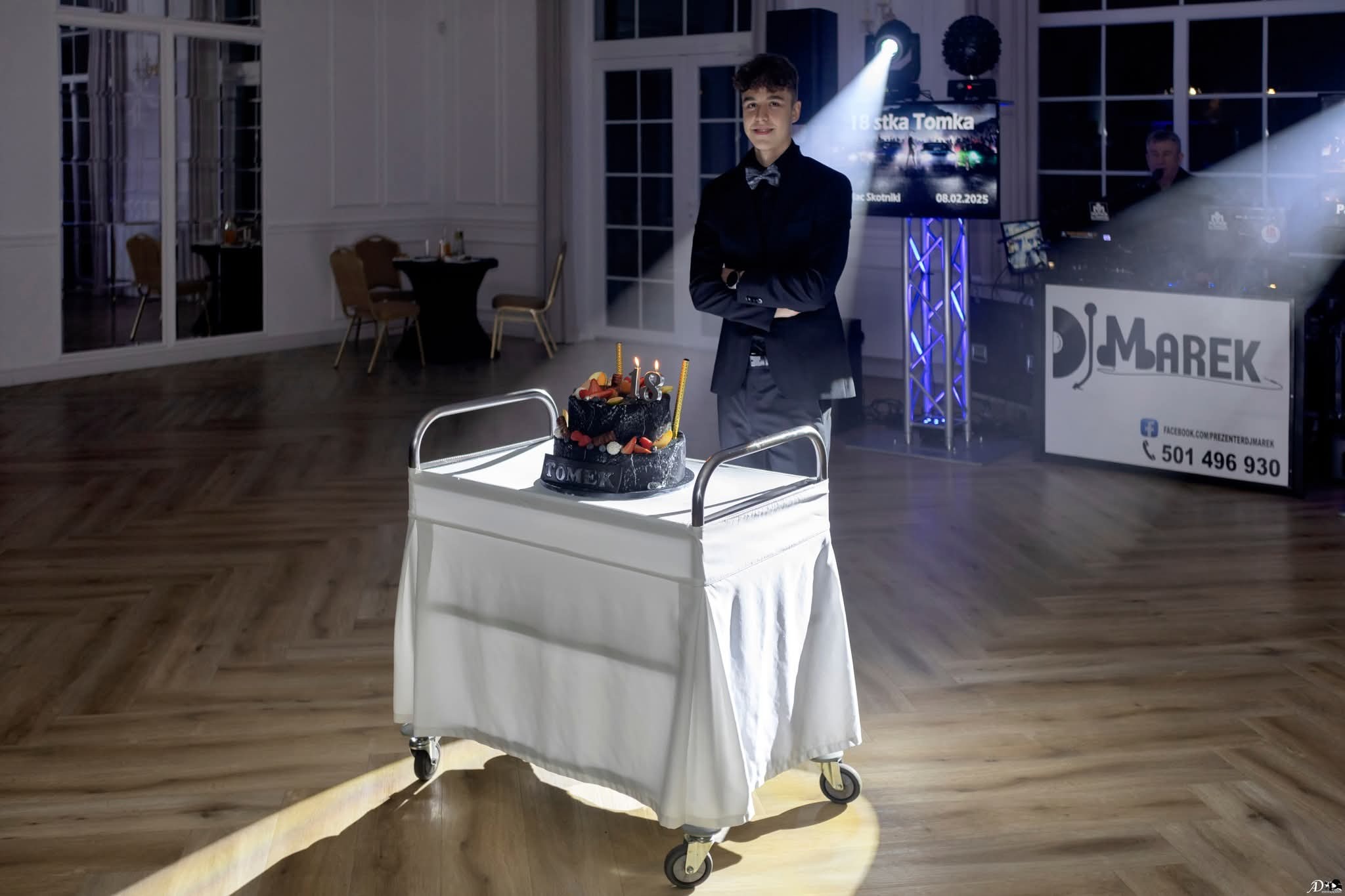 Uśmiechnięty nastolatek w Łodzi na tle sali bankietowej z tortem urodzinowym i stanowiskiem DJ-a, w eleganckim stroju.