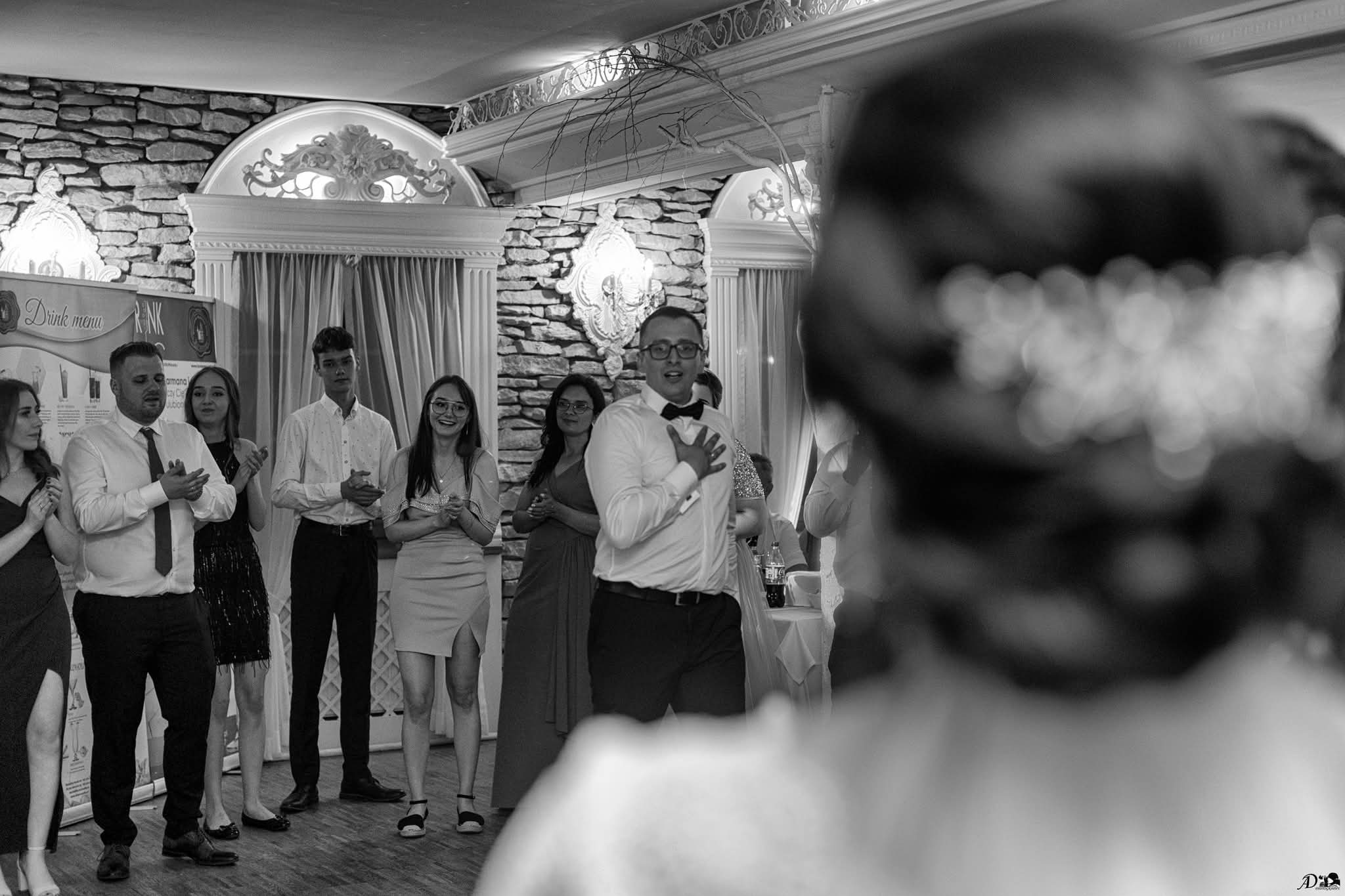 Czarno-białe zdjęcie z wesela: goście weselni, w tym pan młody z muszką, gratulują parze młodej. W tle dekoracje sali i menu drinków.