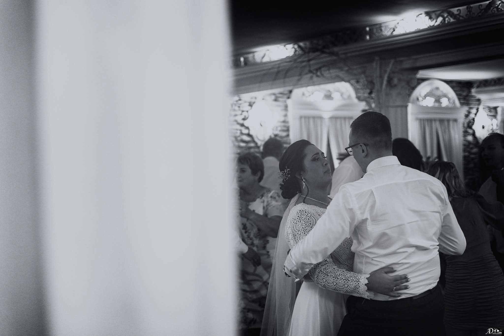 Czarno-białe zdjęcie pary młodej podczas pierwszego tańca na sali weselnej, z widocznymi gośćmi w tle i dekoracyjnym wystrojem wnętrza.