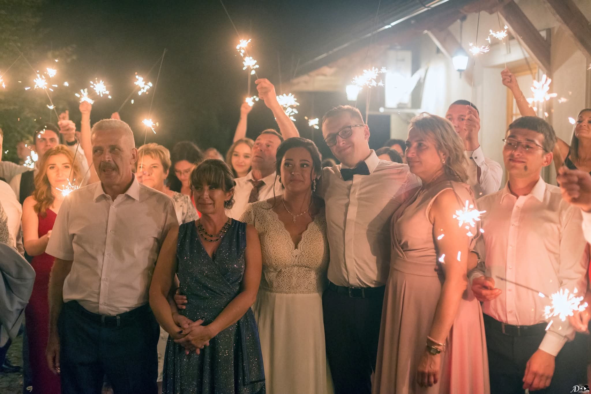 Uśmiechnięta panna młoda w otoczeniu rodziny i gości weselnych, świętujących z zimnymi ogniami w dłoniach. Nocne ujęcie z delikatnym oświetleniem.