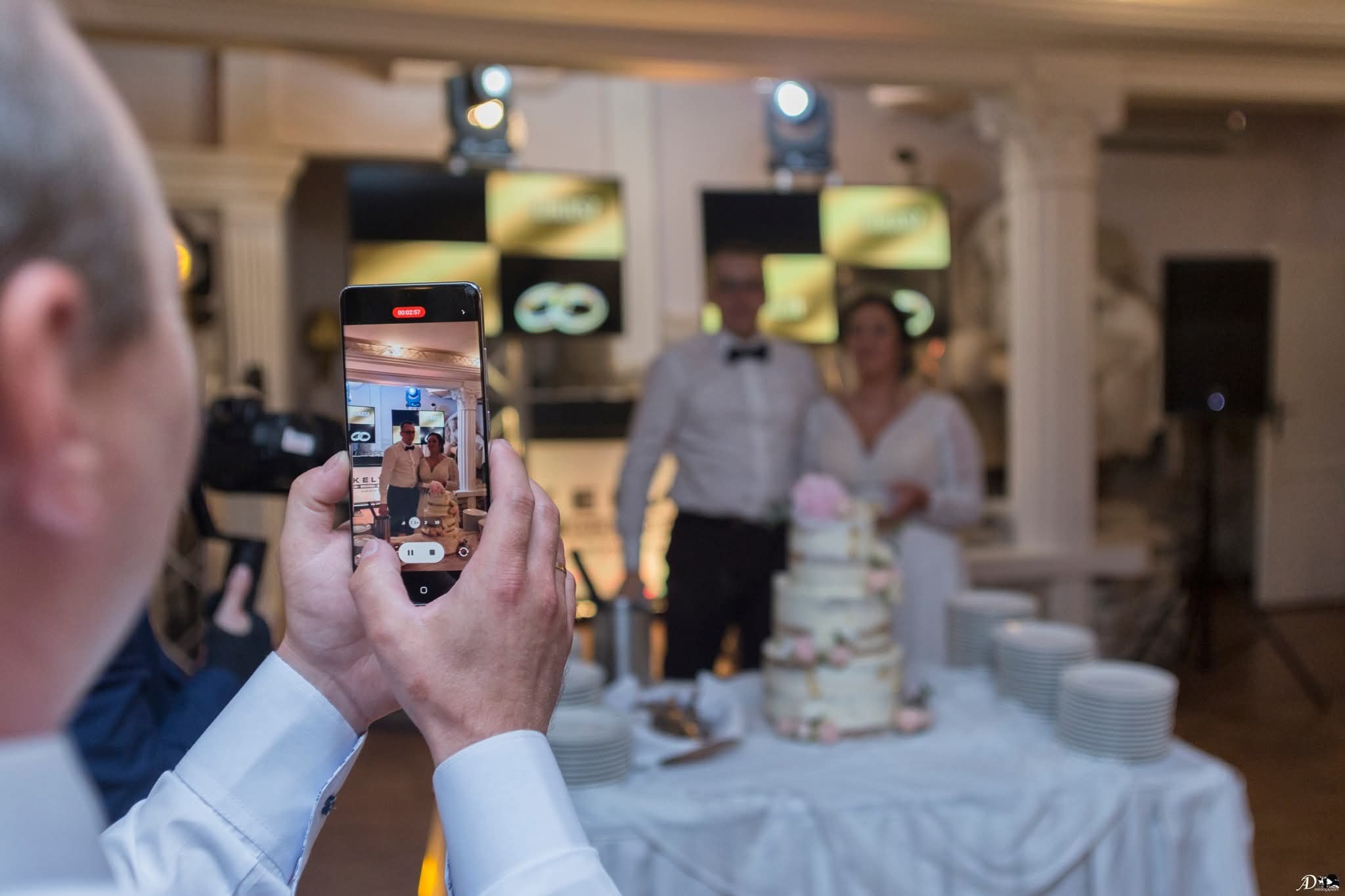 Nagrywanie smartfonem pary młodej i tortu weselnego. Na ekranie telefonu widoczny kadr z wcześniejszego nagrania.