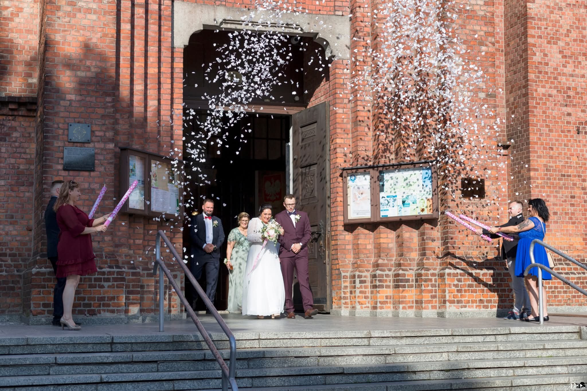 Para młoda wychodzi z kościoła, obsypywana konfetti, ceglana fasada, goście z tubami konfetti, schody wejściowe, jasne światło słoneczne.