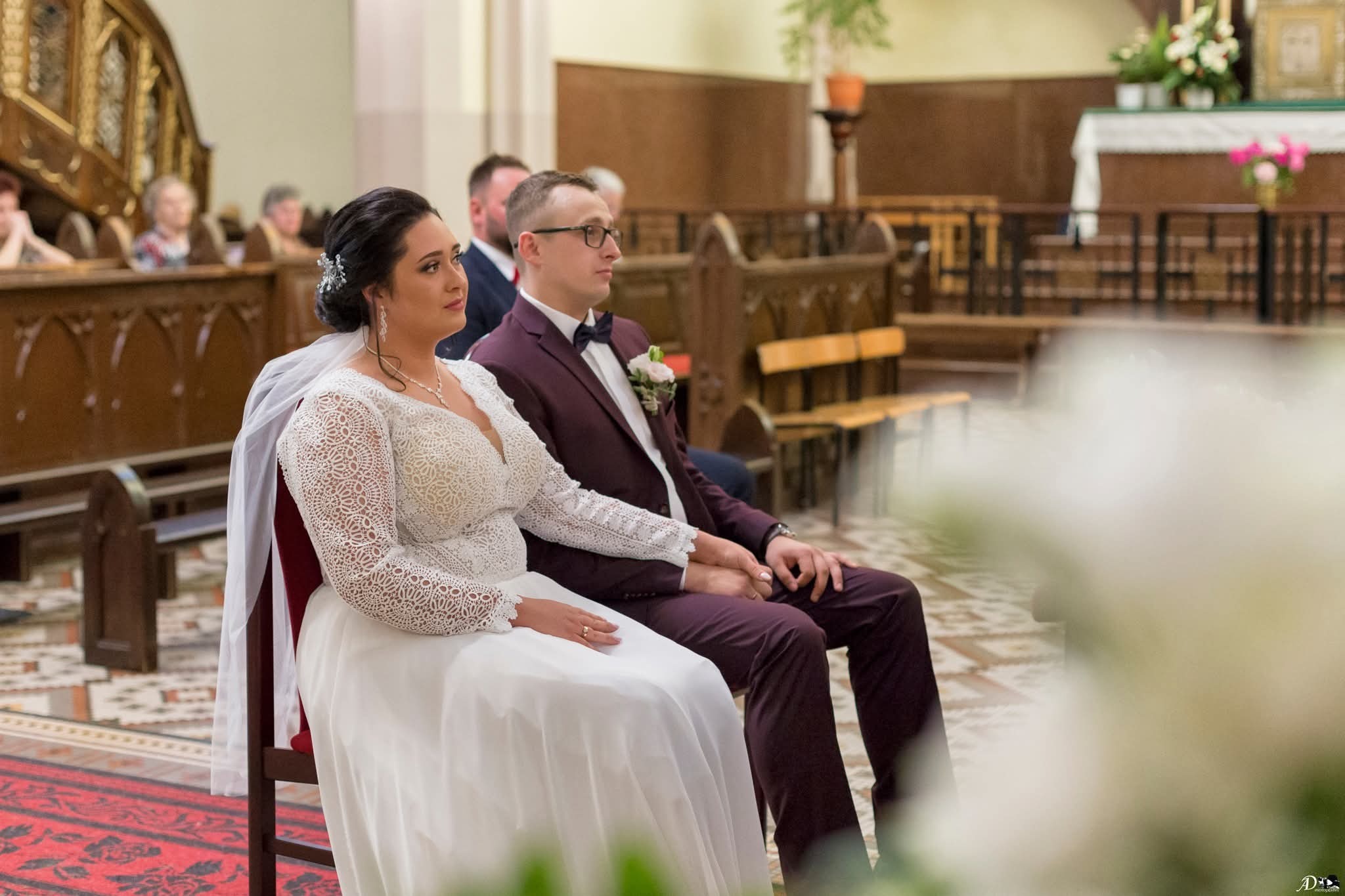 Para młoda siedzi w kościele podczas ceremonii ślubnej. Panna młoda w koronkowej sukni, pan młody w bordowym garniturze. W tle ławki i ołtarz.