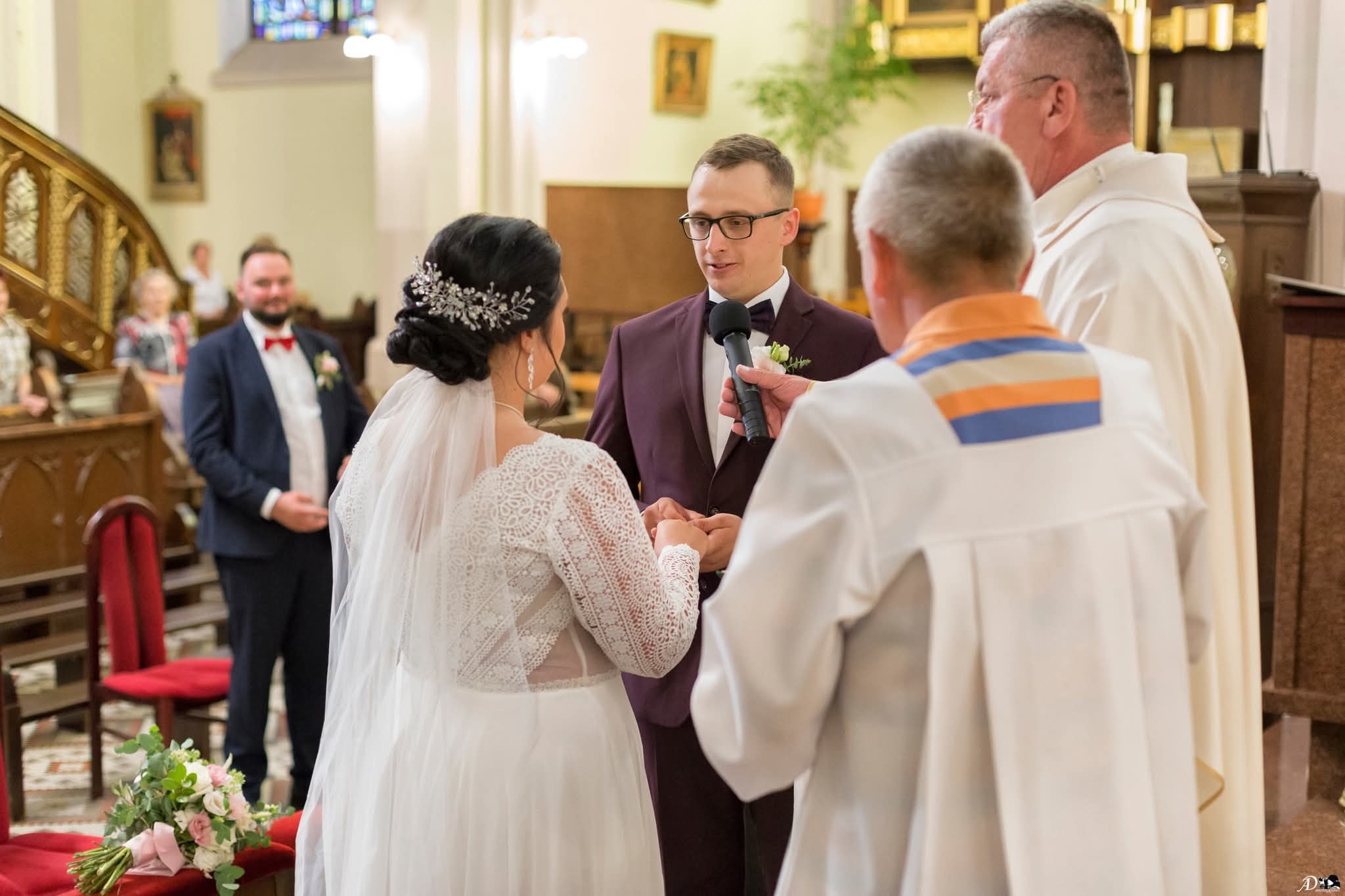 Para młoda w trakcie ceremonii ślubnej w kościele, wymiana obrączek. Pan młody w bordowym garniturze trzyma mikrofon, w tle widoczni goście weselni.