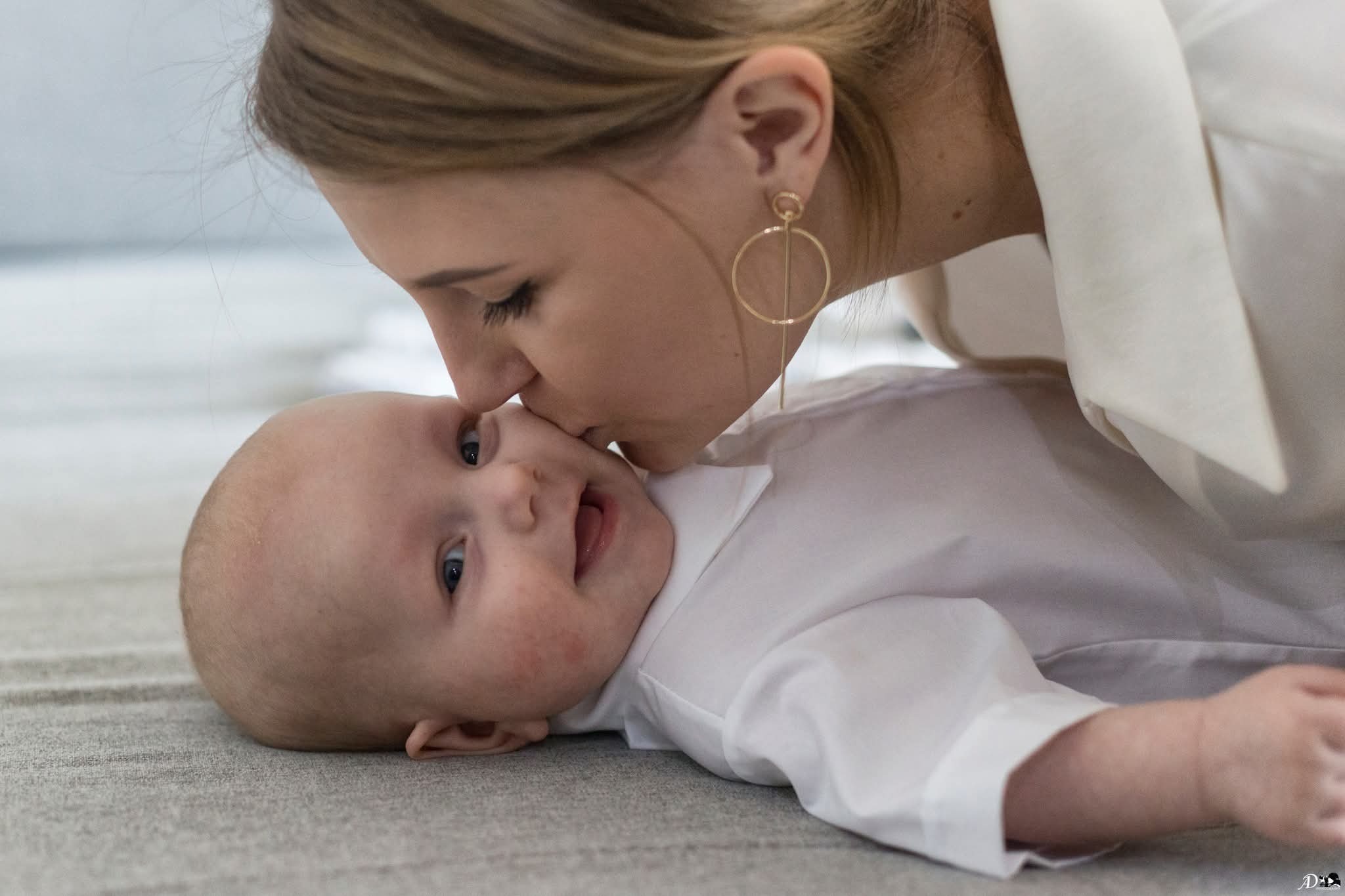 Mama w białej koszuli całuje w czoło leżące na szarym dywanie niemowlę z otwartymi ustami. Dziecko patrzy w górę.