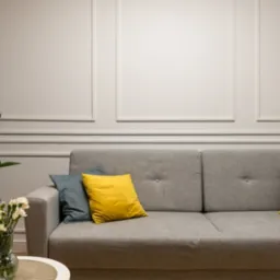 Eleganckie wnętrze salonu z szarą sofą, żółtymi i niebieskimi poduszkami, ścianą z klasyczną sztukaterią, stolikiem z kwiatami i rośliną doniczkową, oświetlone ciepłym światłem.