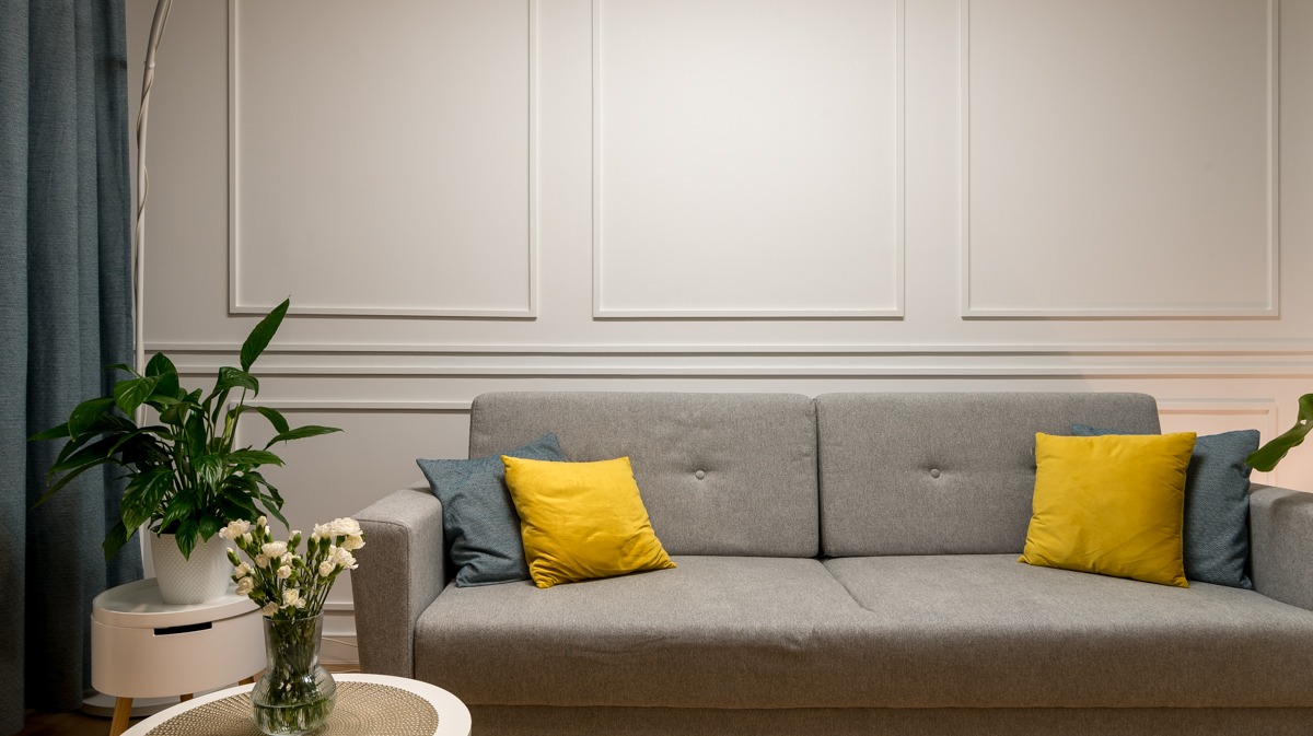 Eleganckie wnętrze salonu z szarą sofą, żółtymi i niebieskimi poduszkami, ścianą z klasyczną sztukaterią, stolikiem z kwiatami i rośliną doniczkową, oświetlone ciepłym światłem.