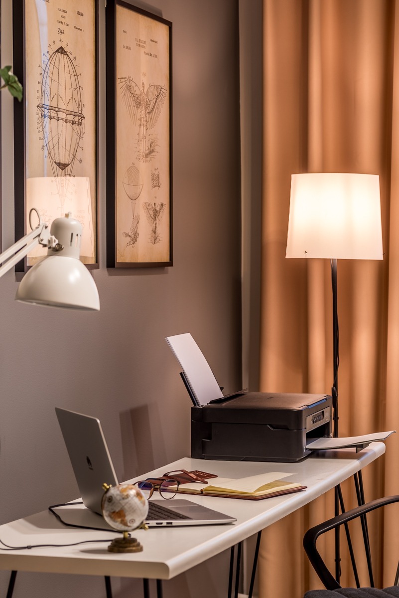 Nowoczesne biuro domowe z białym biurkiem, laptopem, drukarką, lampą podłogową i dekoracyjnymi grafikami w ramkach na szarej ścianie.