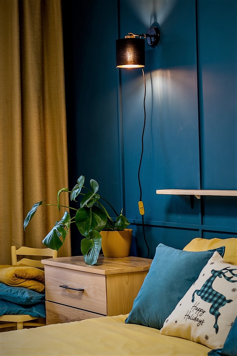 Nastrojowe wnętrze sypialni z ciemnoniebieską ścianą z panelami, drewnianą szafką nocną z rośliną Monstera w żółtej doniczce, czarną lampką ścienną z kablem i żółtym włącznikiem oraz łóżkiem...