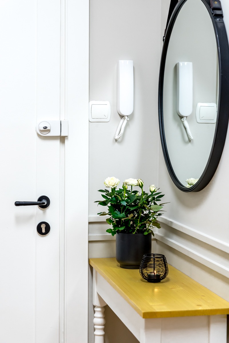 Nowoczesny przedpokój: białe drzwi z czarną klamką, lustro w czarnej ramie, konsola z jasnego drewna z białymi nogami, białe róże w czarnej doniczce i dekoracyjny świecznik.