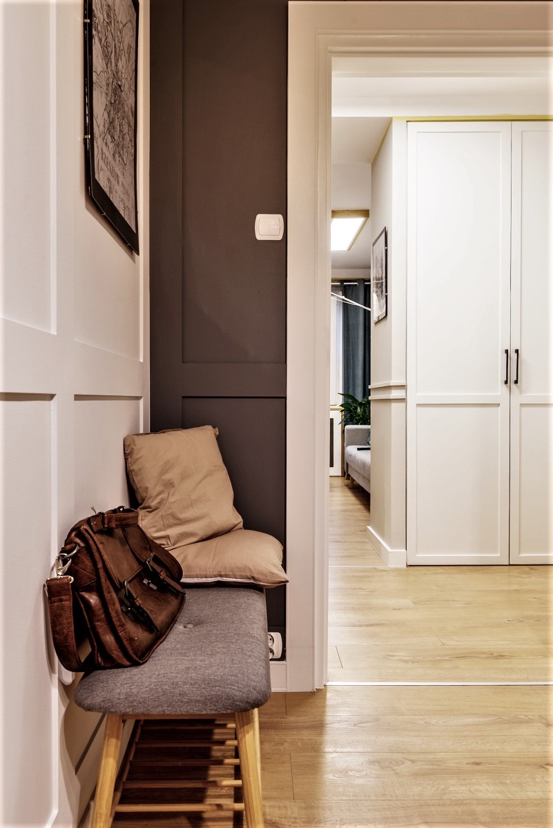 Przedsionek mieszkania z siedziskiem, poduszkami i skórzaną torbą. W tle widoczne drzwi i wnętrze pokoju z drewnianą podłogą.