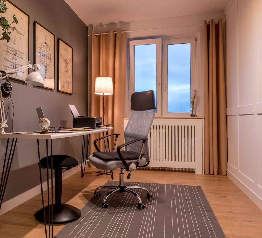 Pokój biurowy z biurkiem na metalowych nogach, ergonomicznym krzesłem, lampą podłogową i dekoracjami ściennymi w odcieniach szarości i beżu.