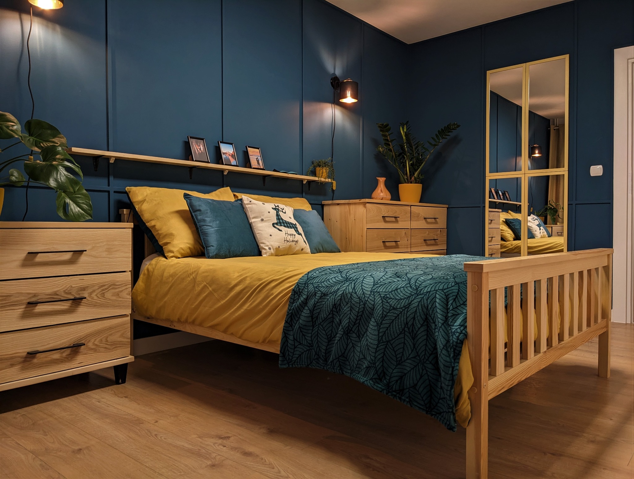 Niebieska sypialnia z drewnianym łóżkiem, komodami i lustrem w złotej ramie. Ściany pomalowane na ciemny niebieski kolor z dekoracyjnymi panelami. Na półce zdjęcia w ramkach i małe rośliny doniczkowe.