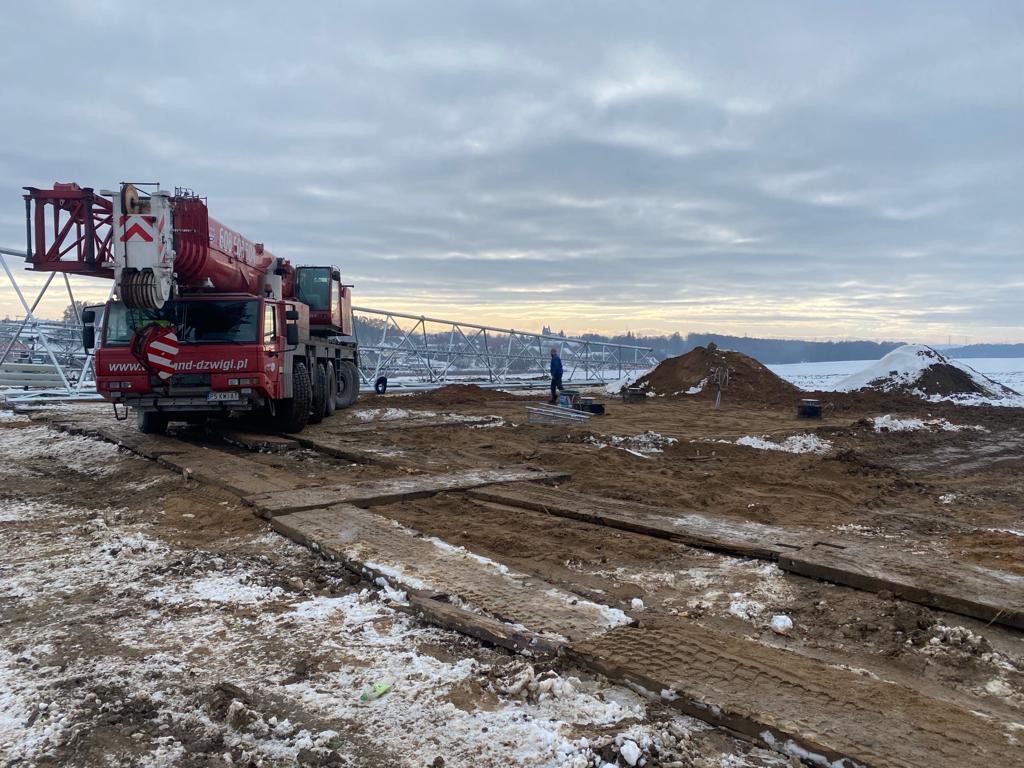 Czerwony dźwig samochodowy na placu budowy drogi, z widoczną konstrukcją mostu w tle i pracownikiem w niebieskiej kurtce, teren częściowo pokryty śniegiem.