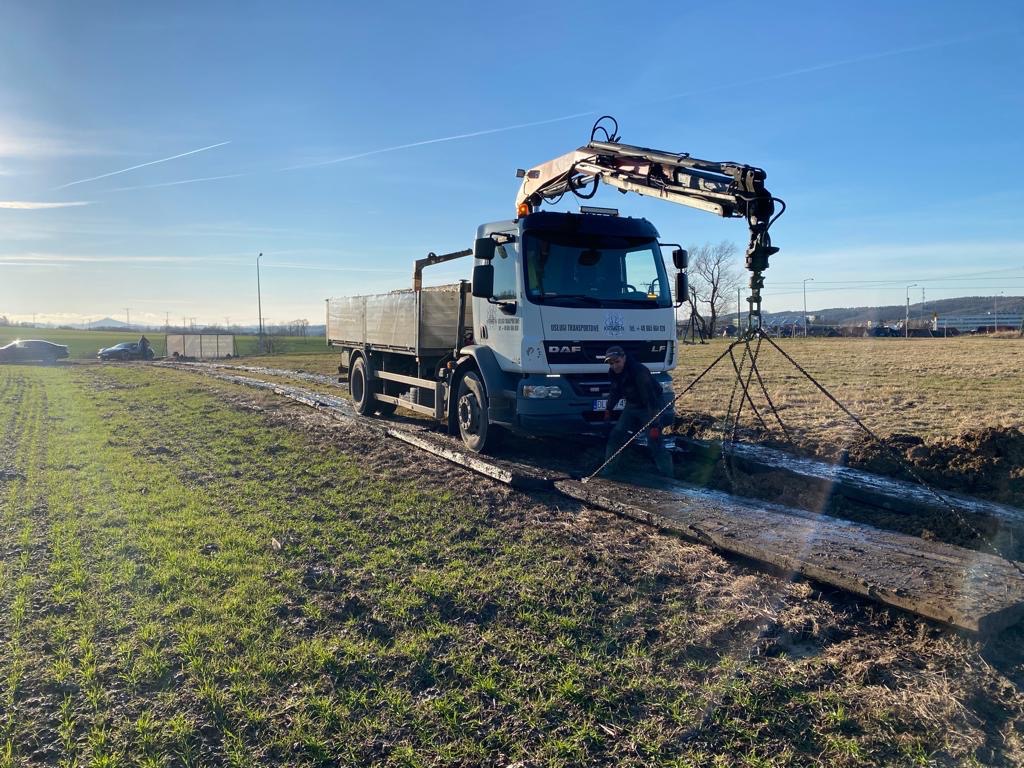 Ciężarówka z dźwigiem podnosi długi, drewniany element na tle zielonego pola i błękitnego nieba. Widoczny pracownik obsługujący dźwig i samochody w oddali.
