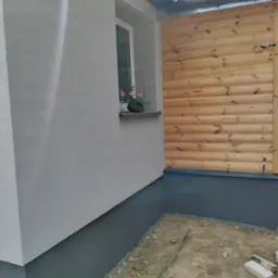 Fragment elewacji domu z oknem, parapetem i doniczką z kwitnącymi różowymi kwiatami, obok drewniana ściana działowa, widoczna szara podmurówka i ziemia.