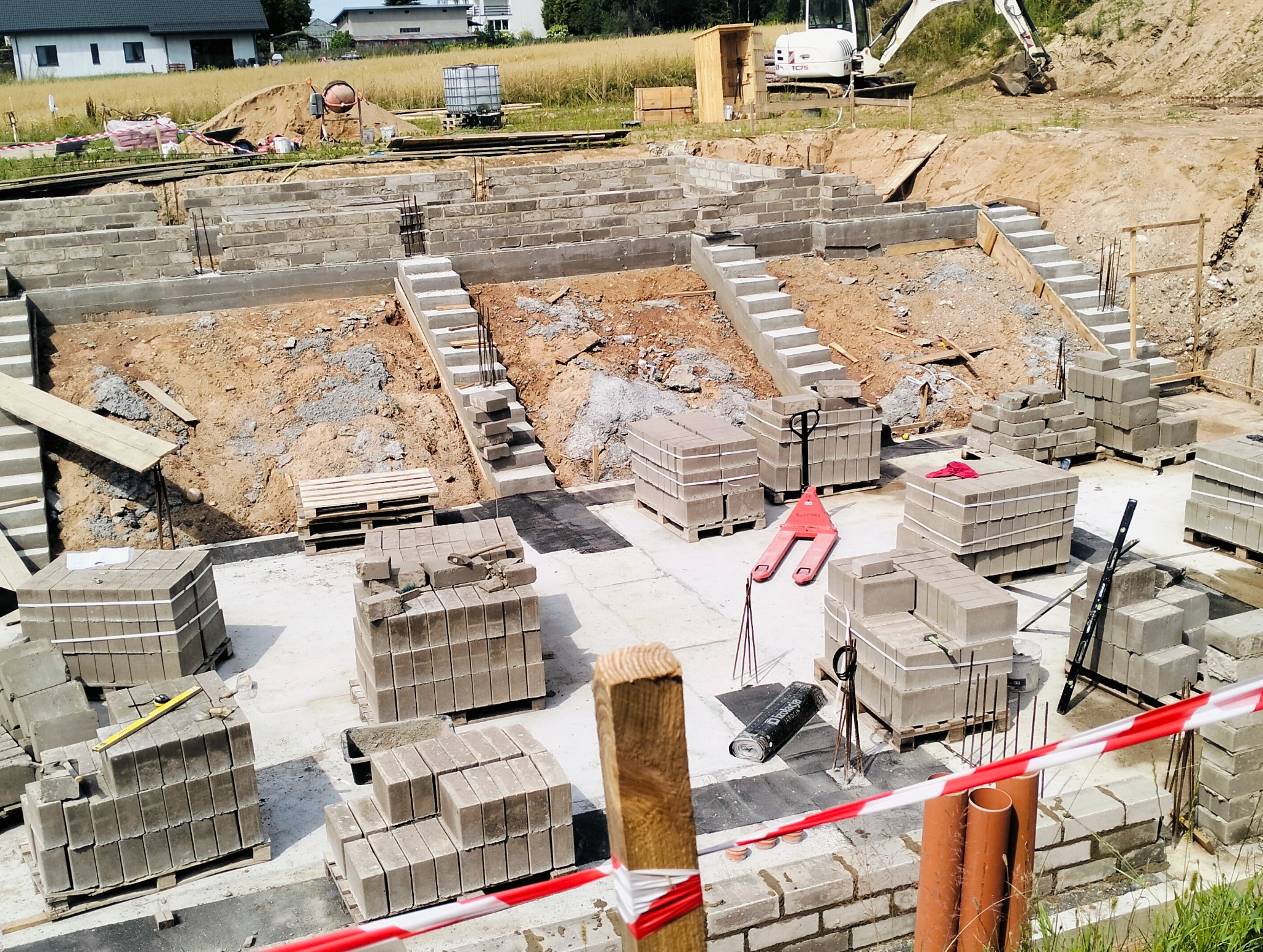 Budowa domu murowanego: widok na fundamenty z bloczków betonowych, schody terenowe, palety z bloczkami, poziomica, taczka, wykop ziemny i koparka w tle.