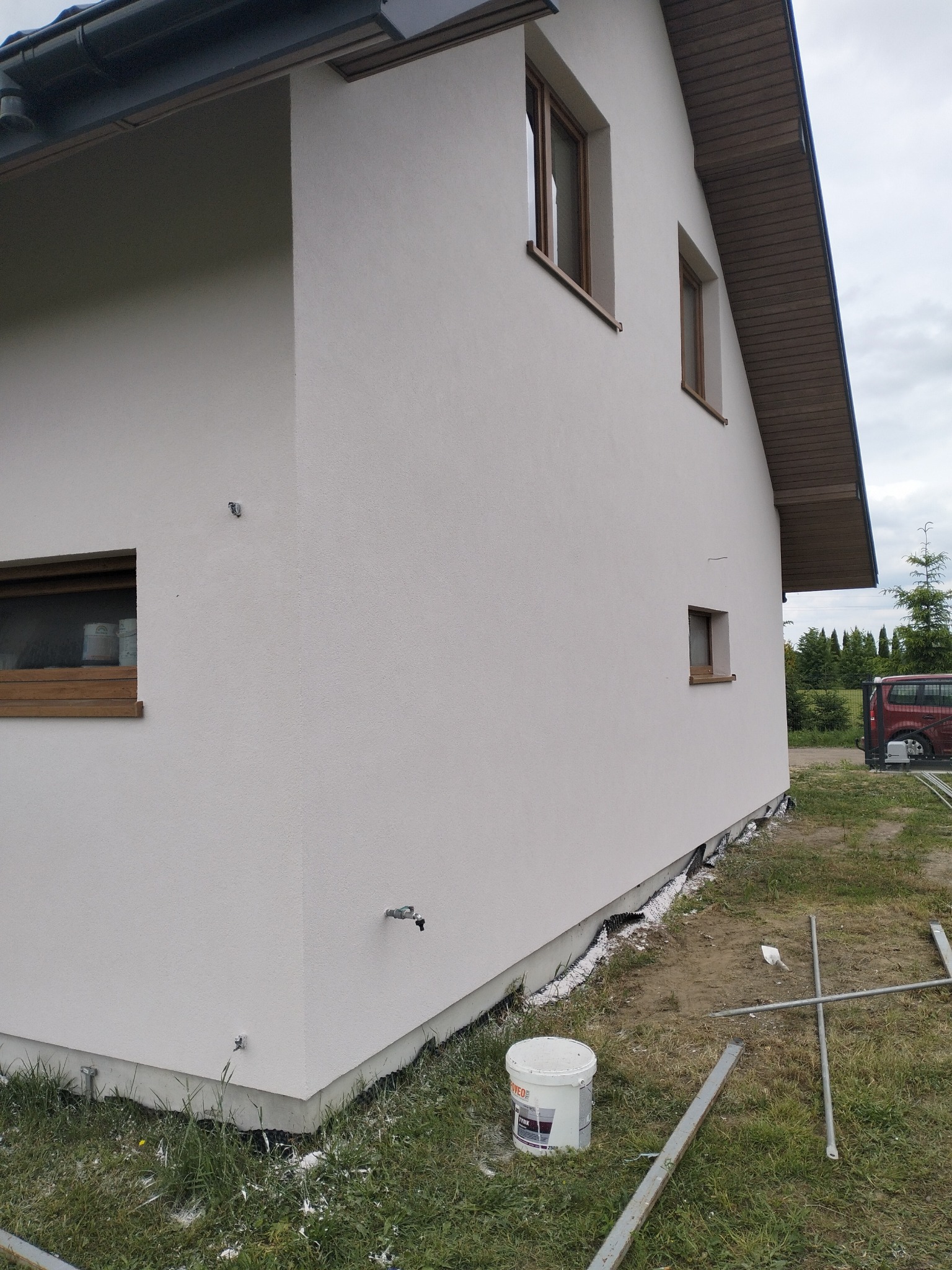 Świeżo otynkowana elewacja domu jednorodzinnego z widocznym fragmentem podbitki dachowej i oknami w drewnianych ramach, z białą farbą rozlaną na trawie i metalowymi elementami rusztowania w pobliżu...